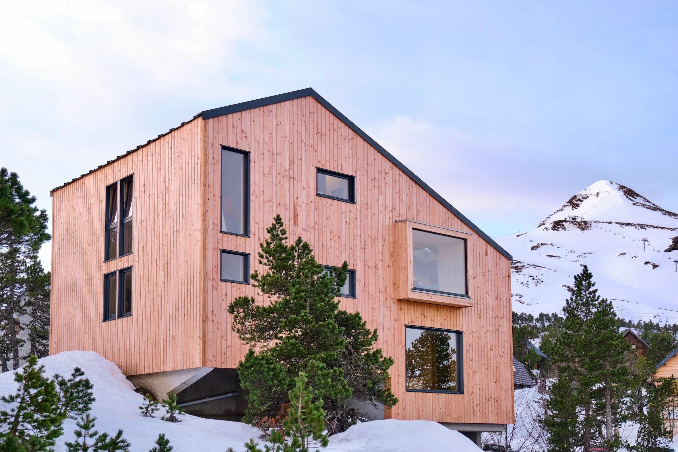 Une maison moderne en bois avec de grandes fenêtres est entourée de neige et de pins. Les sommets des montagnes sont visibles en arrière-plan sous un ciel pâle.