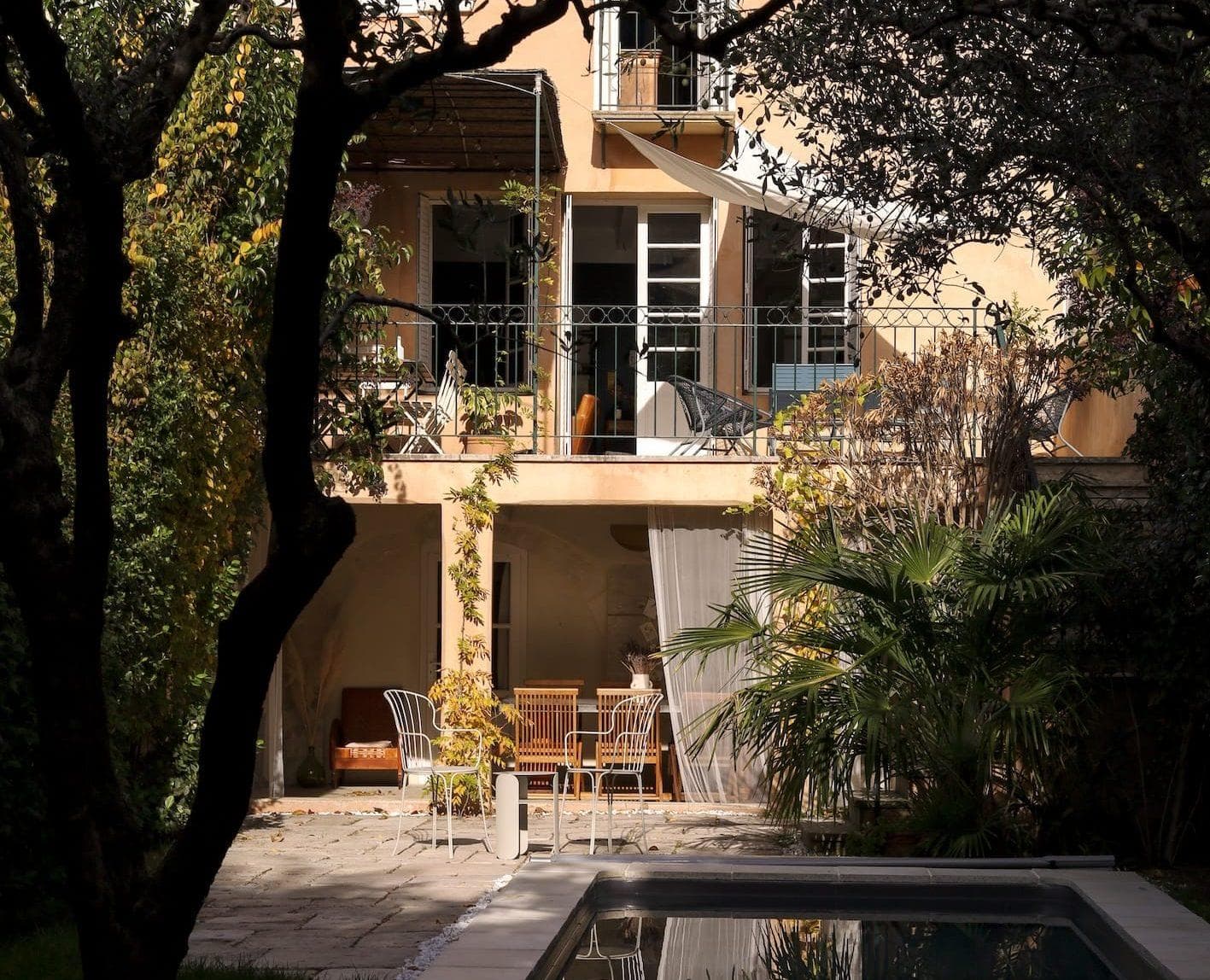 A sunlit backyard with a patio of a two-story house. The patio has tables and chairs surrounded by trees and plants. There’s a swimming pool in the foreground, and the house has balconies with additional seating.