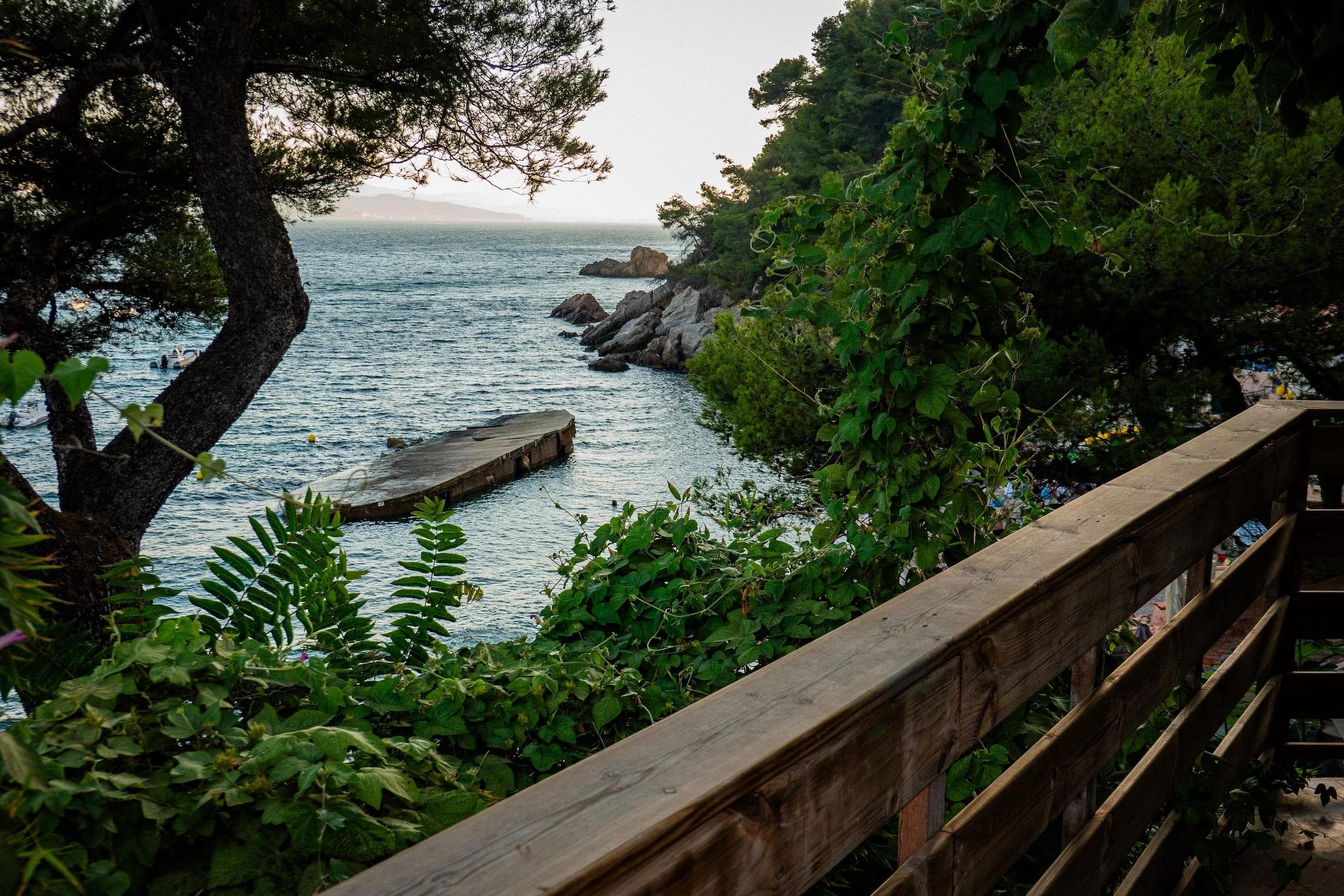 Une vue d'une rambarde en bois surplombant une mer calme. Une dense végétation verte entoure la scène, et une plateforme en béton s'étend dans l'eau. Des arbres encadrent l'image, avec un horizon dégagé visible en arrière-plan.