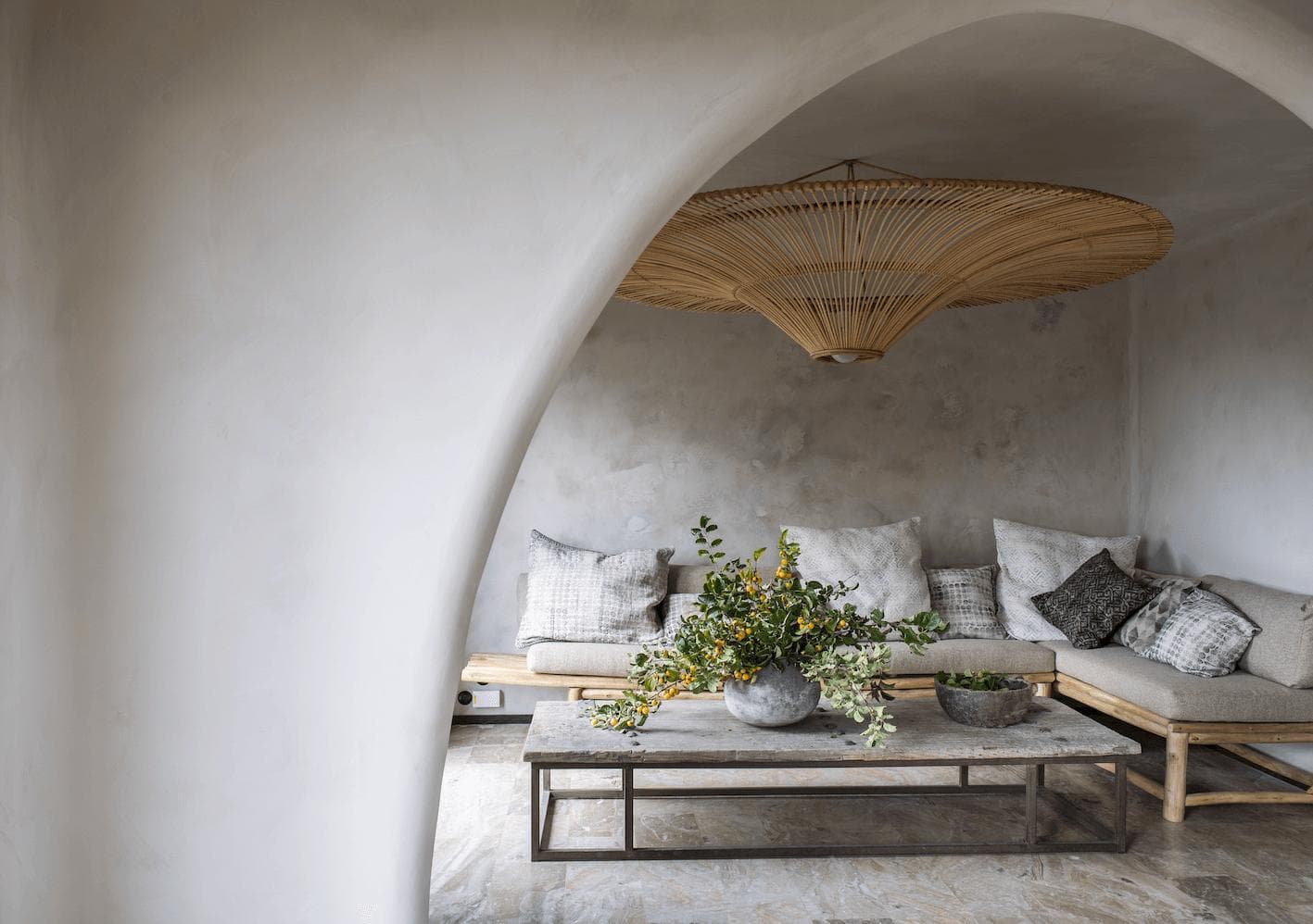 Modern living room with a minimalist design featuring a large, decorative hanging light fixture. The space includes a comfortable sectional sofa with cushions, a wooden coffee table displaying a vase with greenery, and an arched entryway.