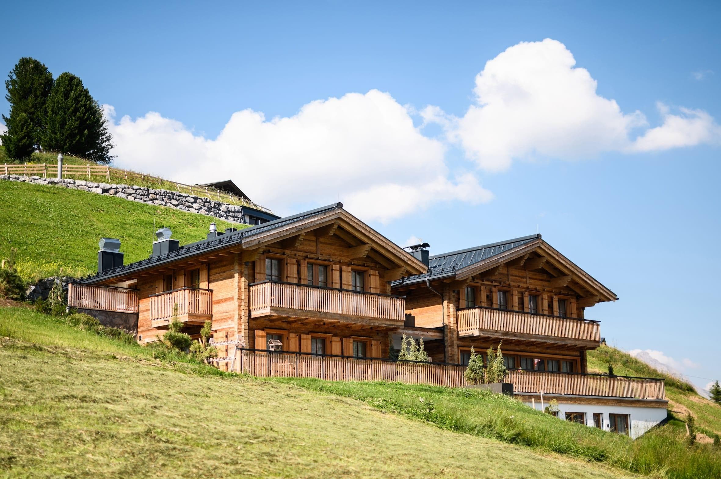 Deux chalets en bois sur une colline herbeuse sous un ciel bleu parsemé de nuages cotonneux. Quelques arbres et un mur de pierre sont visibles en arrière-plan, renforçant le cadre tranquille et pastoral.