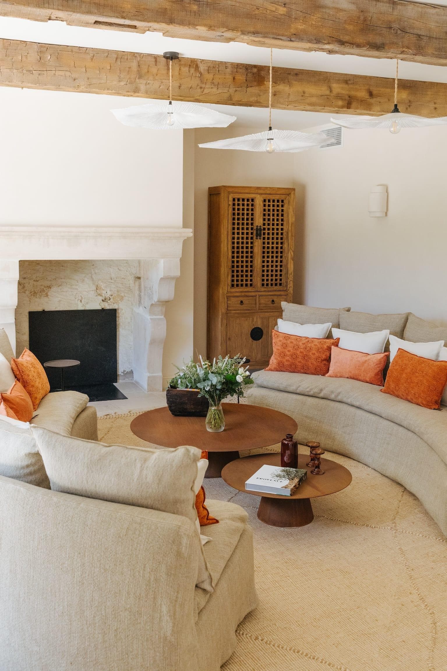 A cozy living room with beige sofas and orange cushions, a round wooden coffee table with flowers, a stone fireplace, wooden ceiling beams, and a tall wooden cabinet against a cream-colored wall.