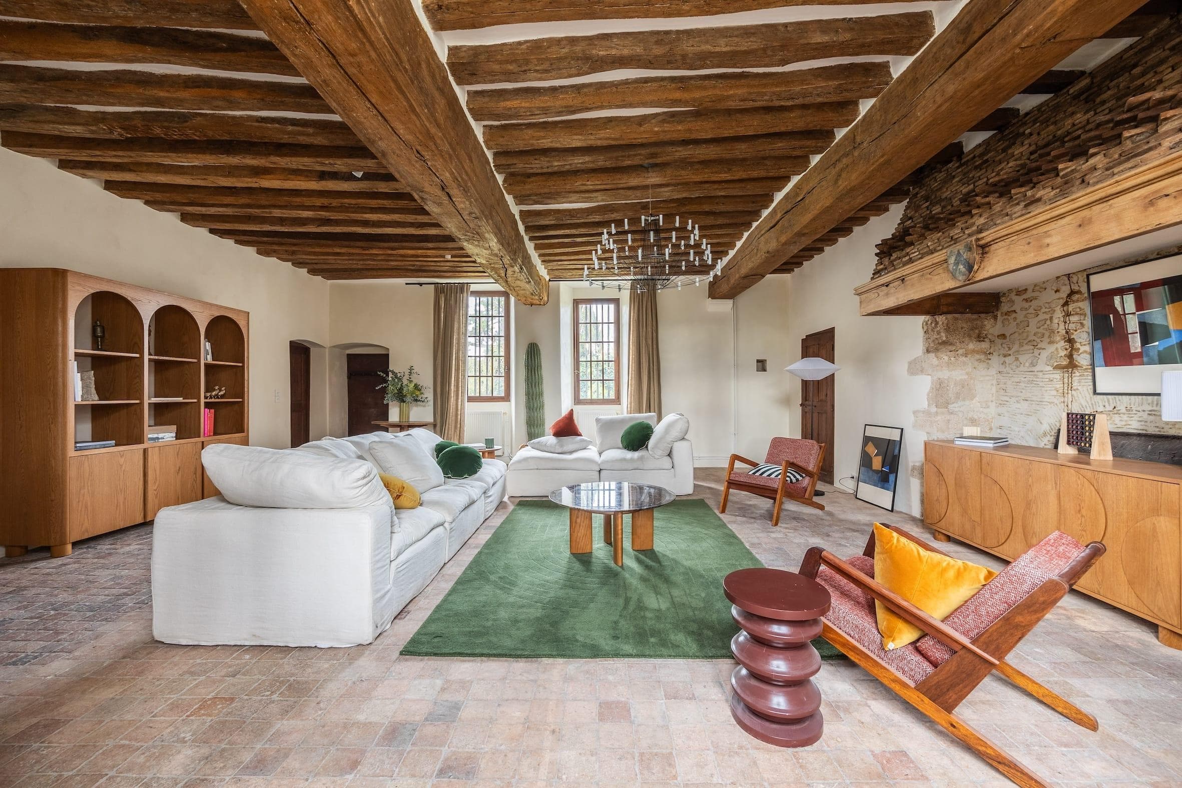 Un salon rustique avec poutres apparentes et murs en pierre comprend des canapés blancs, un tapis vert, des meubles en bois et des coussins colorés. Deux grandes fenêtres laissent entrer la lumière naturelle et un lustre moderne est suspendu au plafond.