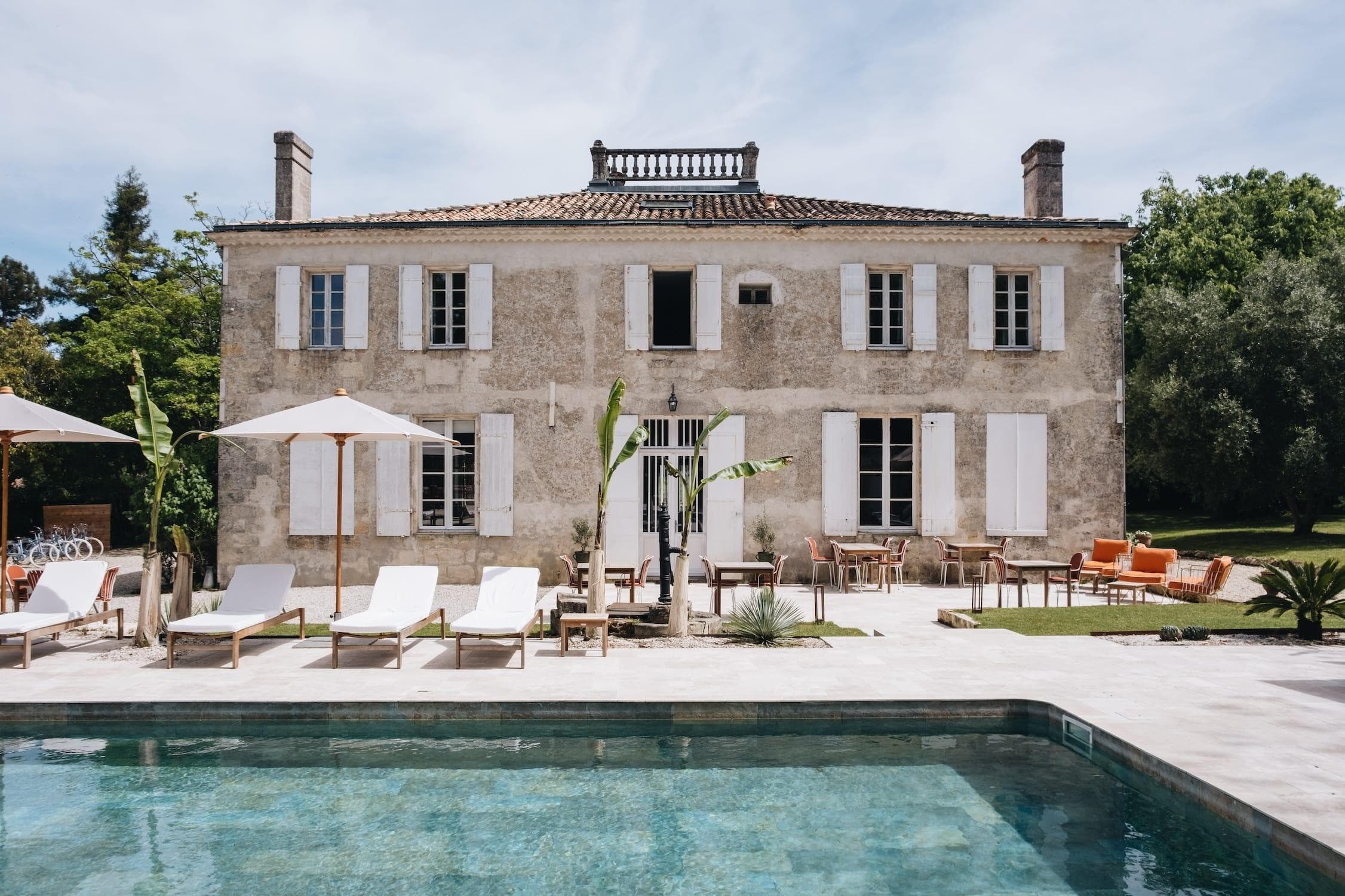 A rustic two-story stone villa with white shutters, surrounded by greenery, features a swimming pool in the foreground. Lounge chairs and umbrellas are arranged by the pool, and outdoor seating is visible on the patio.