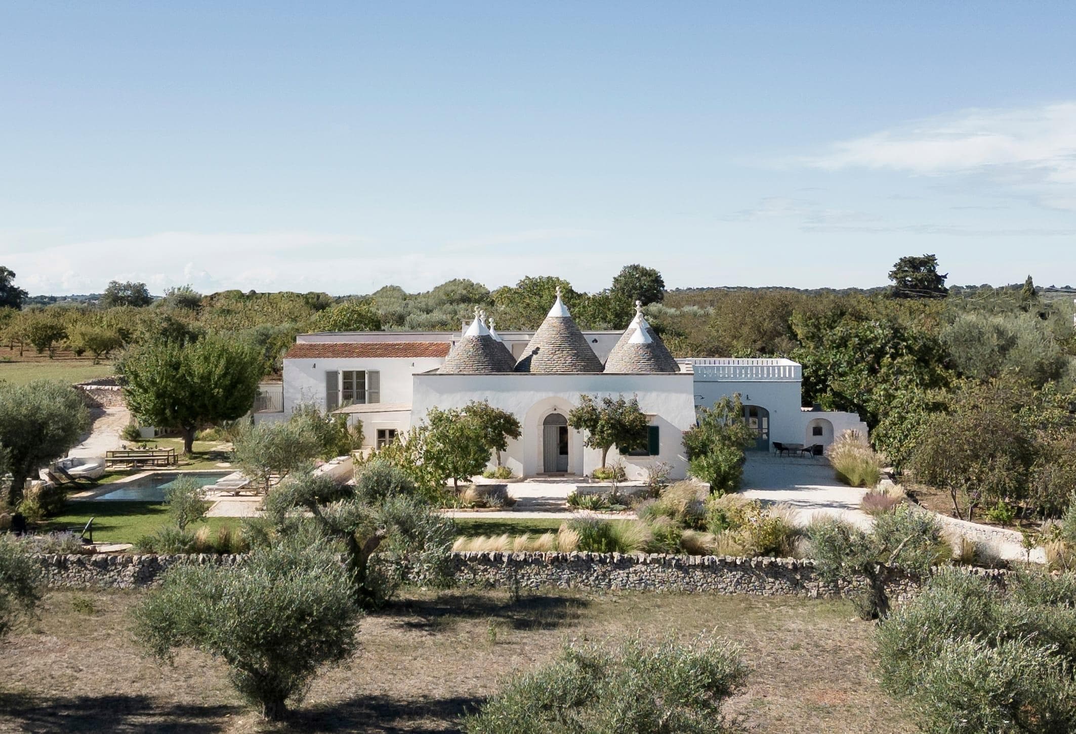 Un bâtiment blanc aux toits coniques se dresse dans un paysage rural entouré d'oliviers et de champs. L'architecture est traditionnelle, avec un mur de pierre rustique et une petite piscine dans le jardin. Le ciel est clair et bleu.