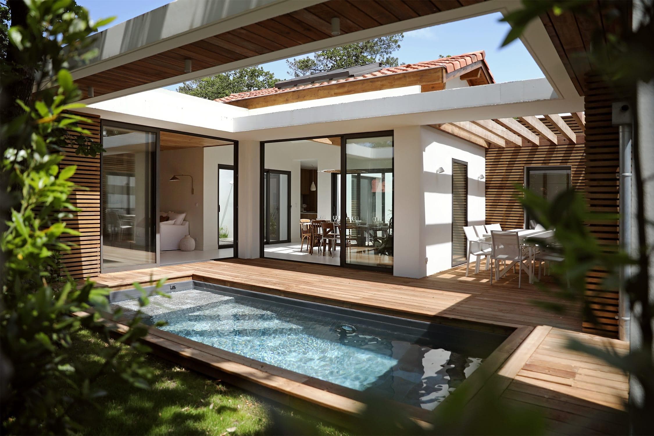 Maison moderne avec cour intérieure dotée d'une petite piscine rectangulaire entourée d'une terrasse en bois. De grandes portes vitrées relient les espaces intérieurs et extérieurs. Des sièges et des plantes d'extérieur renforcent l'atmosphère sereine.