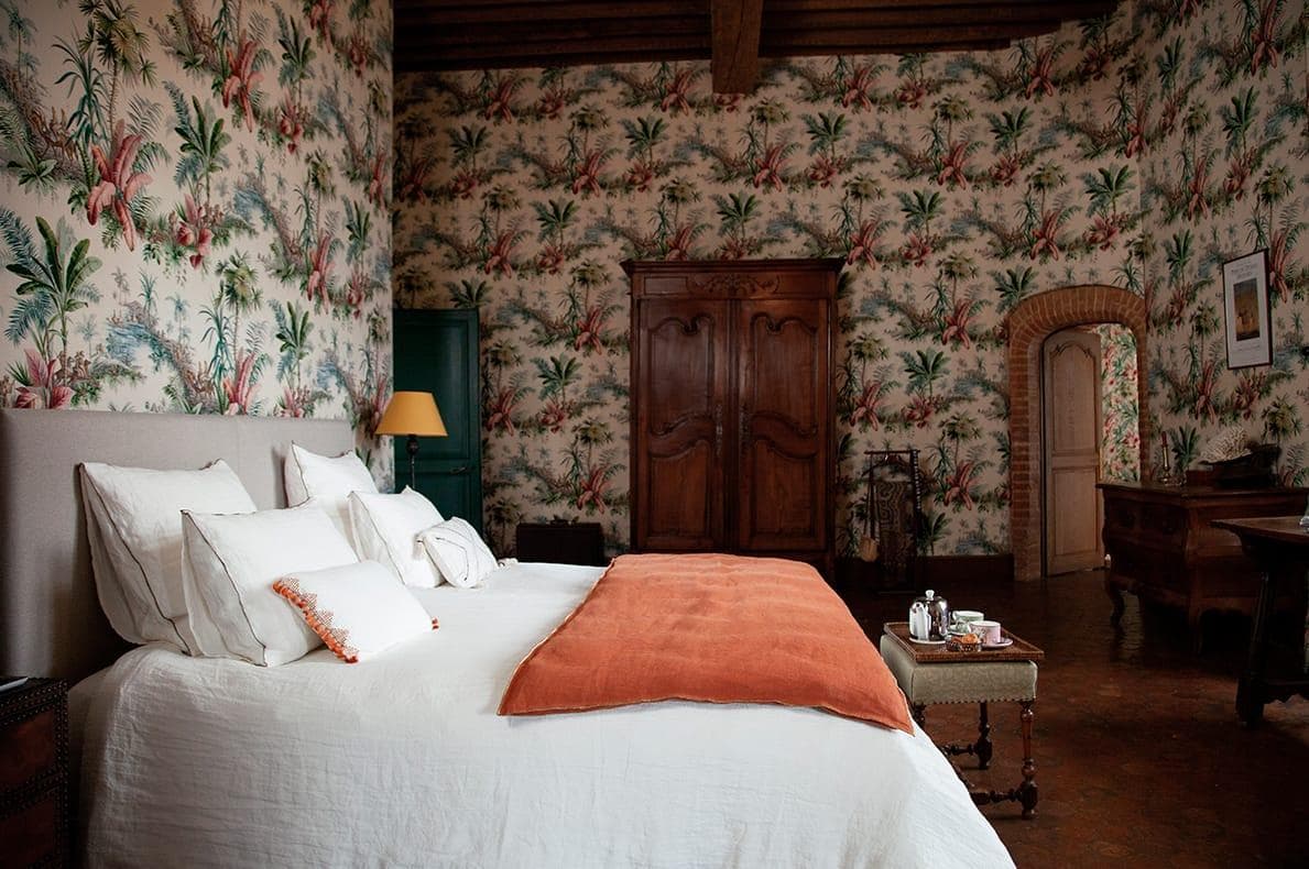 A comfortable room with tropical floral wallpaper, including a large bed with white sheets and an orange plaid. There's a wooden wardrobe, a table with a lamp and a tray with cups. The room exudes a rustic yet elegant atmosphere.