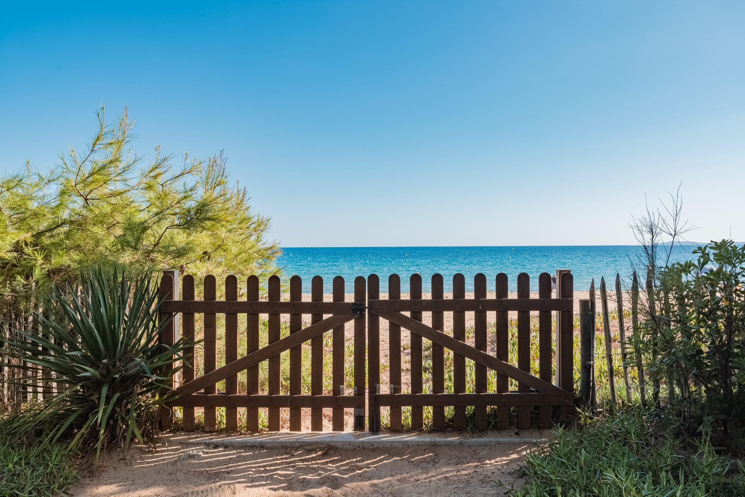 Un portail en bois s'ouvre sur une plage de sable, offrant à la maison une vue pittoresque avec accès direct. Encadré par une végétation verte luxuriante des deux côtés, le ciel bleu clair rencontre l'horizon serein de l'océan, créant une scène côtière paisible.