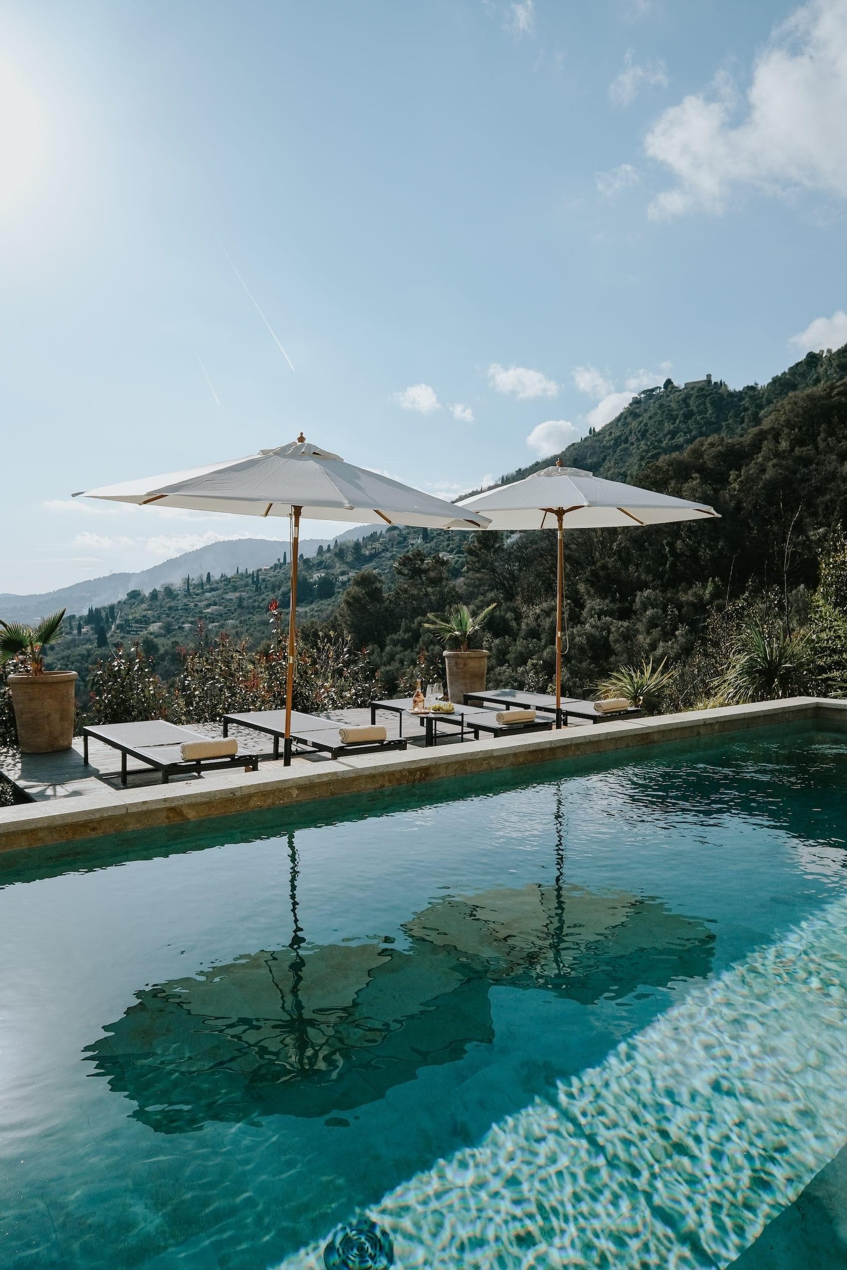 Une piscine extérieure sereine avec deux chaises longues et des parasols blancs se trouve au milieu d'une verdure luxuriante et de collines sous un ciel bleu clair.