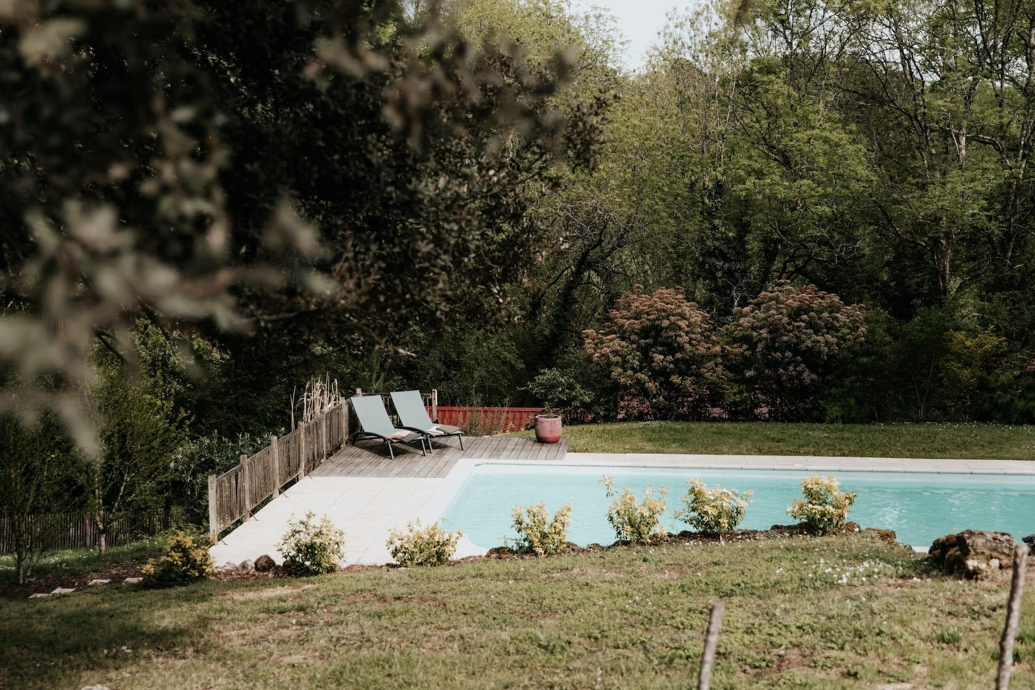 Une scène extérieure tranquille avec une piscine entourée d'une végétation luxuriante et d'arbres. Deux chaises longues sont placées sur la terrasse au bord de la piscine et une jardinière rose se trouve à proximité. Le cadre est serein et accueillant.