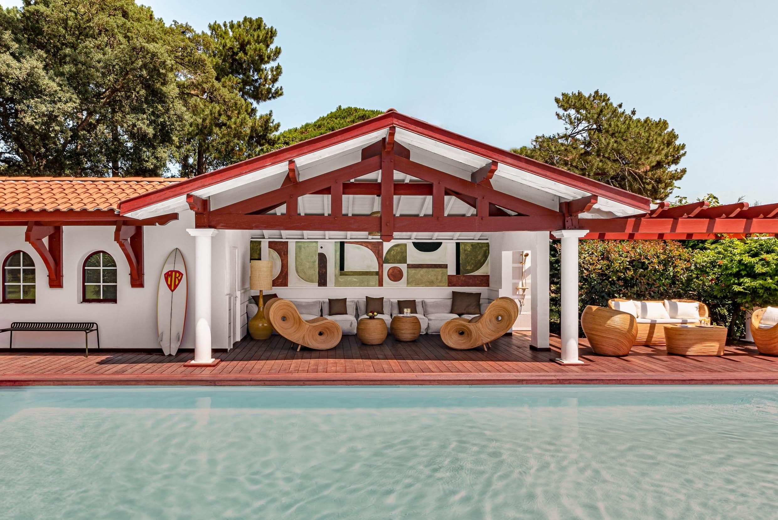 A stylish poolside lounge area at a location villa avec piscine Hossegor, featuring wicker furniture under a red and white wooden pavilion, modern wall art, a surfboard, and lush green trees in the background. The scene is bright and inviting.