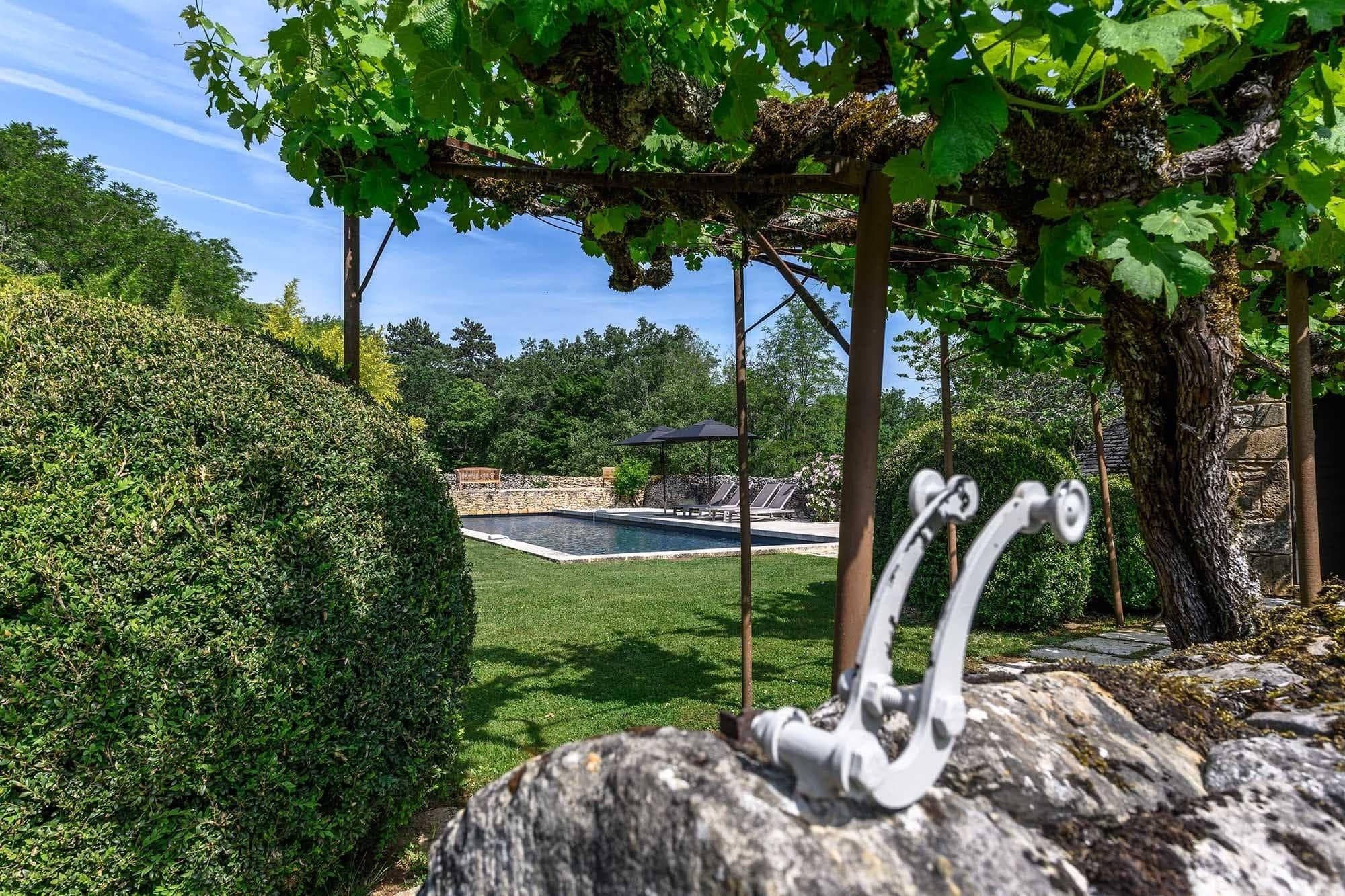 Un jardin luxuriant avec des buissons taillés et une pergola mène à une piscine rectangulaire en arrière-plan. Une pièce décorative en métal est placée au premier plan sur une surface en pierre. Des arbres et un ciel bleu complètent le cadre tranquille.