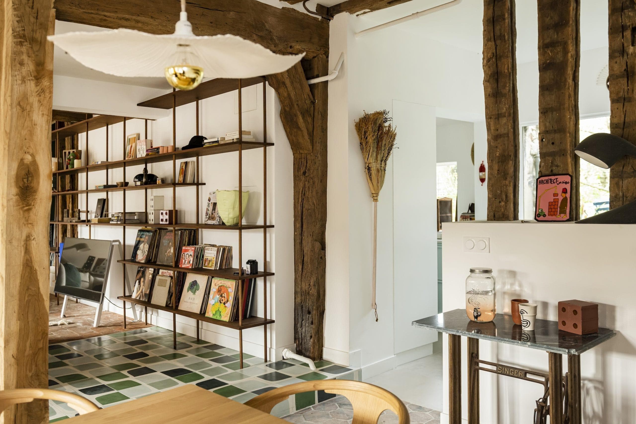 Une pièce chaleureuse et rustique avec des poutres en bois apparentes et des étagères modernes remplies de livres et d'œuvres d'art. Un sol carrelé vert et blanc complète les accents en bois. Un balai est accroché au mur près d'une petite table avec des objets décoratifs et une bougie en pot.
