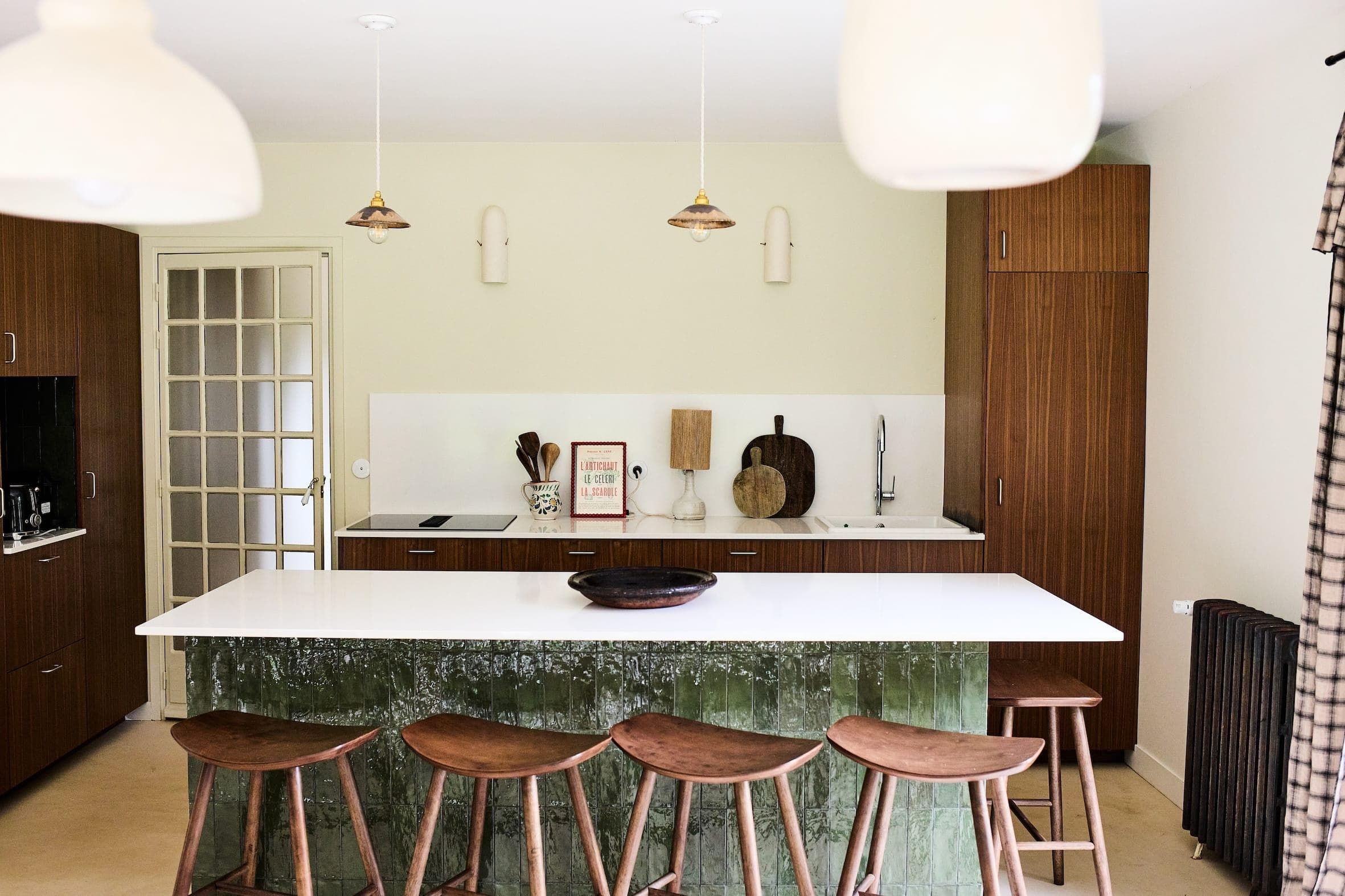 Une cuisine moderne avec des armoires en bois et un grand îlot blanc. Cinq tabourets en bois bordent l'îlot. Des luminaires suspendus illuminent l'espace. Une porte vitrée s'ouvre sur le côté, tandis que des ustensiles de cuisine et des objets de décoration ornent le comptoir.