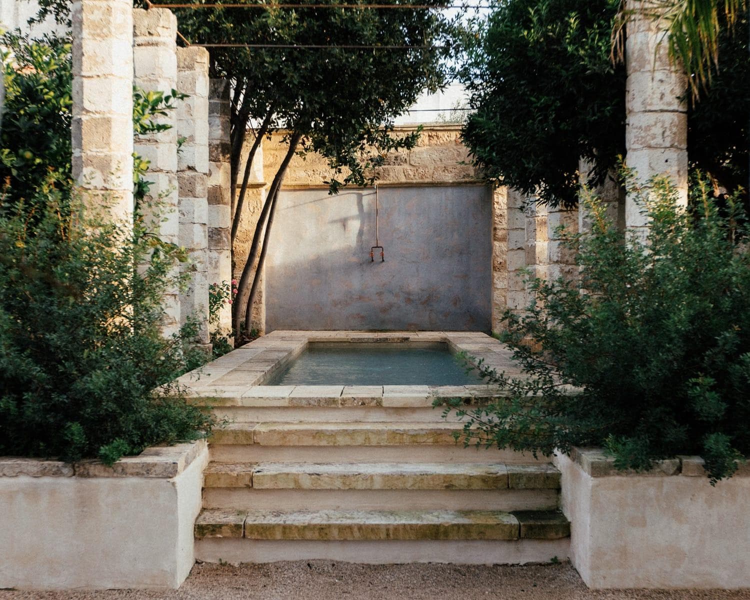 Stone path leading to a small rectangular pool surrounded by stone columns and greenery. Steps lead down to the pool, with trees and a stone wall in the background, creating a serene, rustic ambience.