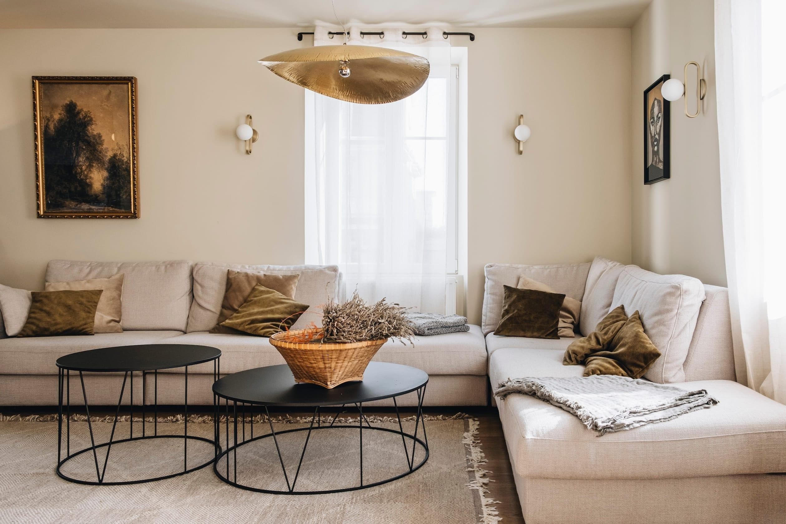 A cozy living room features a beige sectional sofa with green and brown cushions. Two round black coffee tables hold a wicker basket of dried flowers. Light filters through sheer curtains, and decorative wall art adds a touch of elegance.