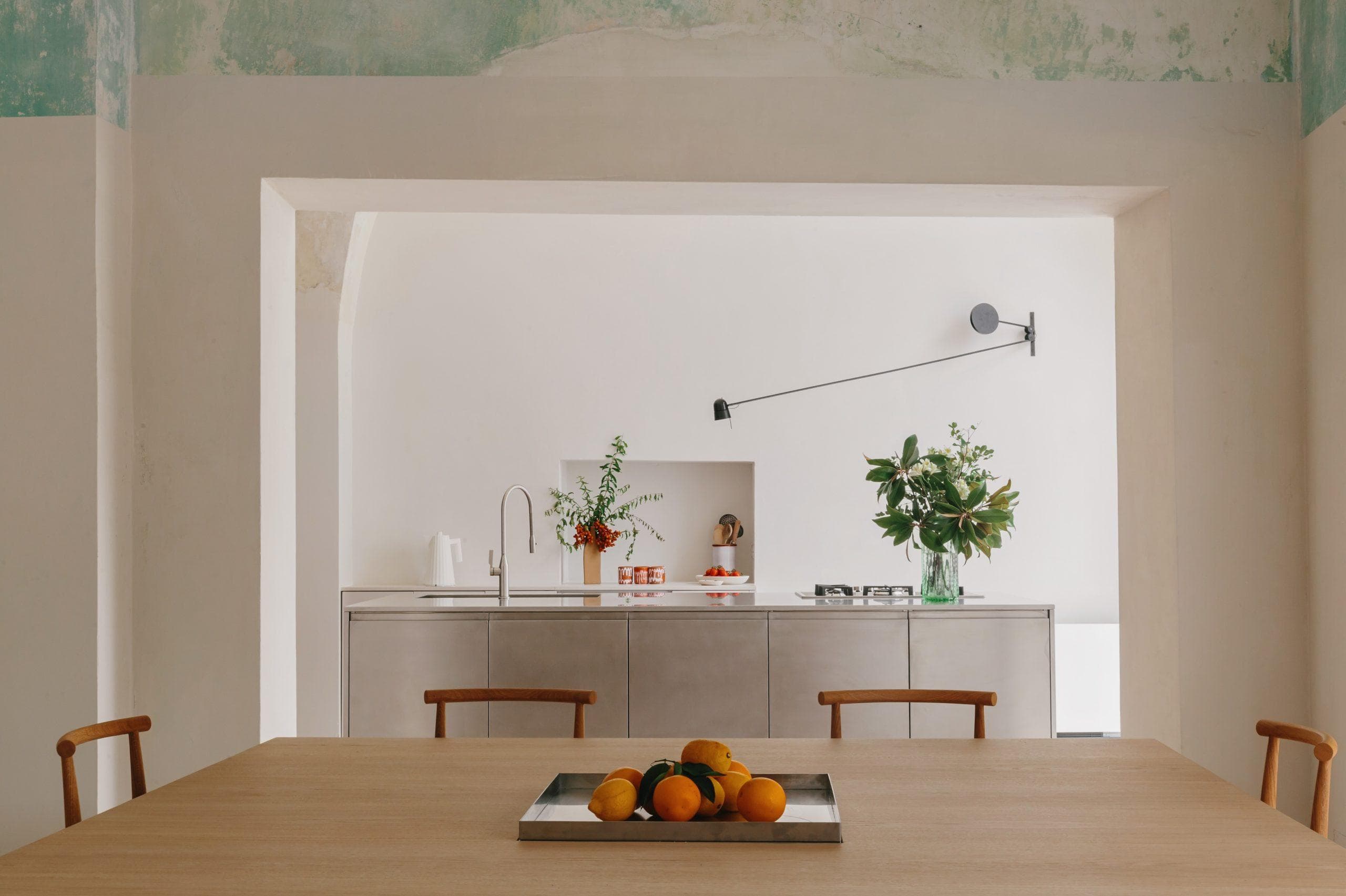 Une cuisine minimaliste comprend une table à manger en bois clair avec un plateau en métal rempli d'oranges au premier plan. Des armoires en acier inoxydable, une applique murale et des vases avec de la verdure et des fleurs sont visibles sur un fond de murs blancs avec de la peinture verte écaillée.