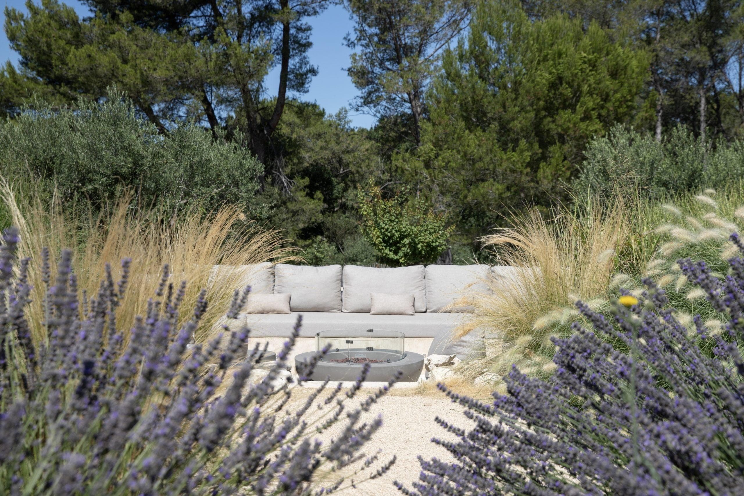Un coin salon extérieur paisible entouré de plantes de lavande éclatantes et de diverses herbes. Un canapé gris avec coussins se trouve derrière un brasero circulaire moderne, avec en toile de fond des arbres verdoyants sous un ciel bleu clair.