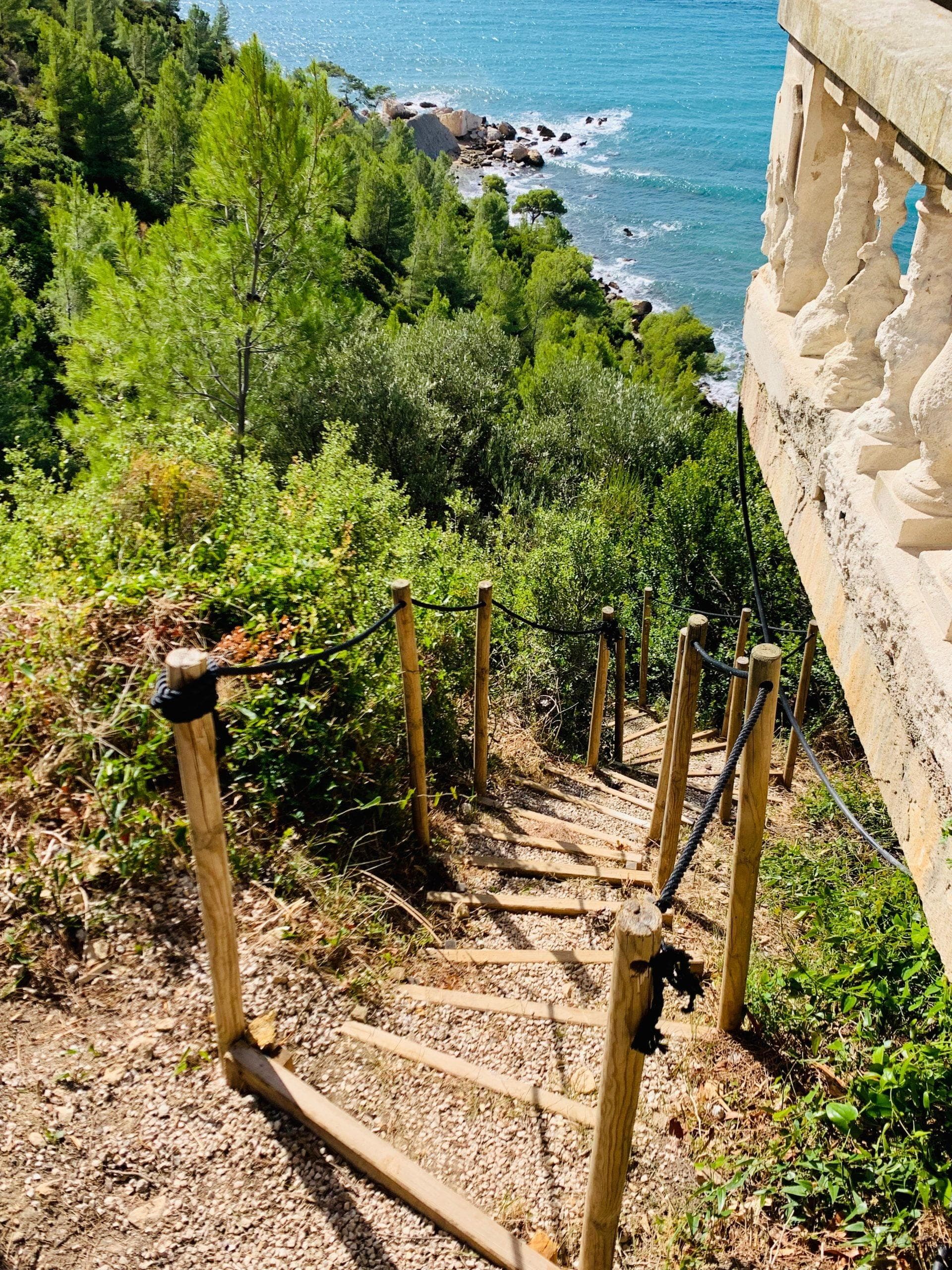 Un étroit escalier en bois descend abruptement sur une colline herbeuse, bordée d'une balustrade en pierre. En arrière-plan, la vue panoramique de cet endroit luxueux révèle la mer d'un bleu éclatant qui rencontre un littoral rocheux sur la Côte d'Azur, le tout encadré par des arbres verts luxuriants.