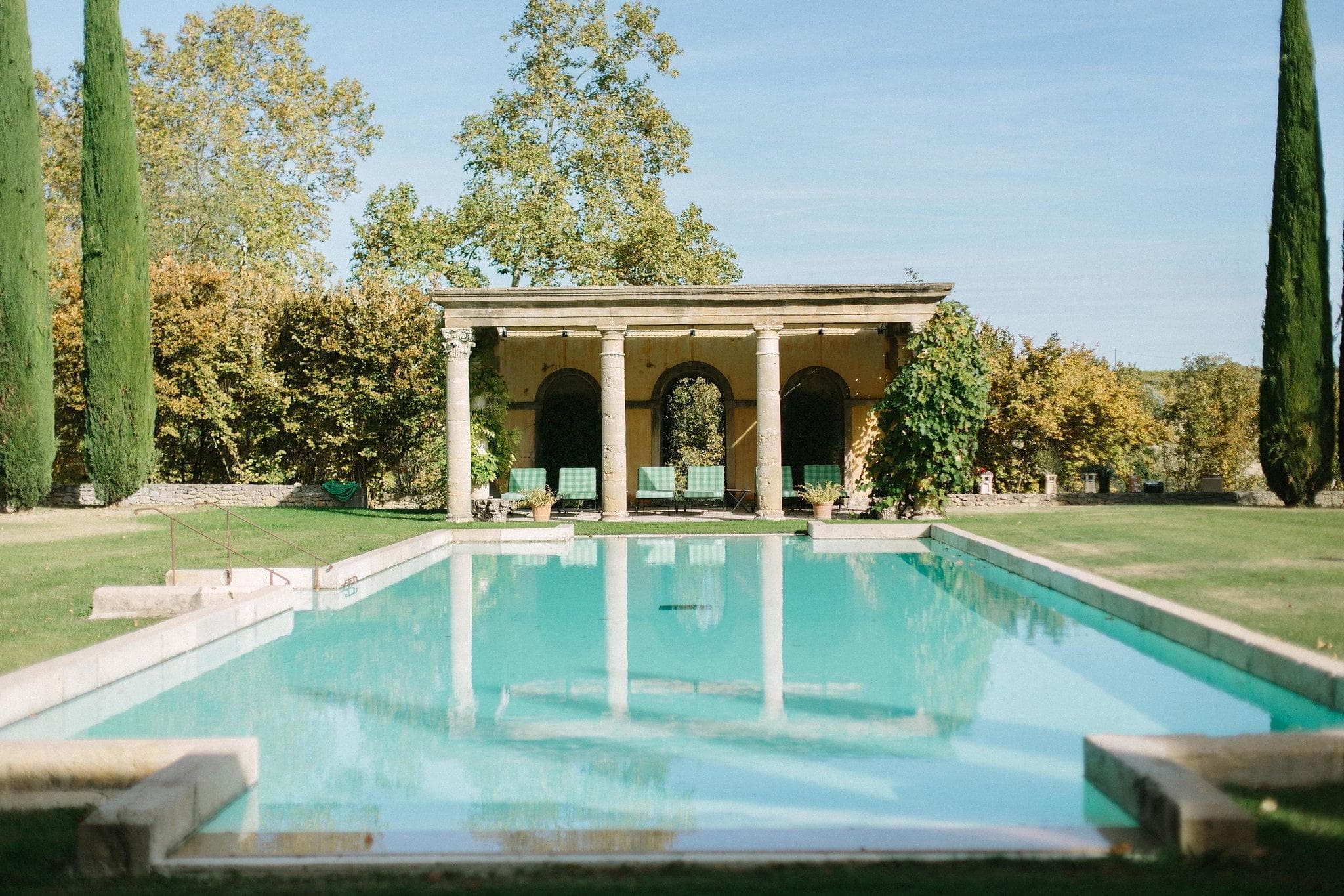 La sereine piscine rectangulaire du Château de Sannes - l'une des meilleures locations de vacances de luxe en France - est bordée de pelouses vertes et de grands cyprès. À l'extrémité, un pavillon en pierre à trois arches se dresse, offrant un espace ombragé pour s'asseoir. Le ciel bleu clair et la végétation luxuriante se reflètent dans l'eau calme de la piscine.