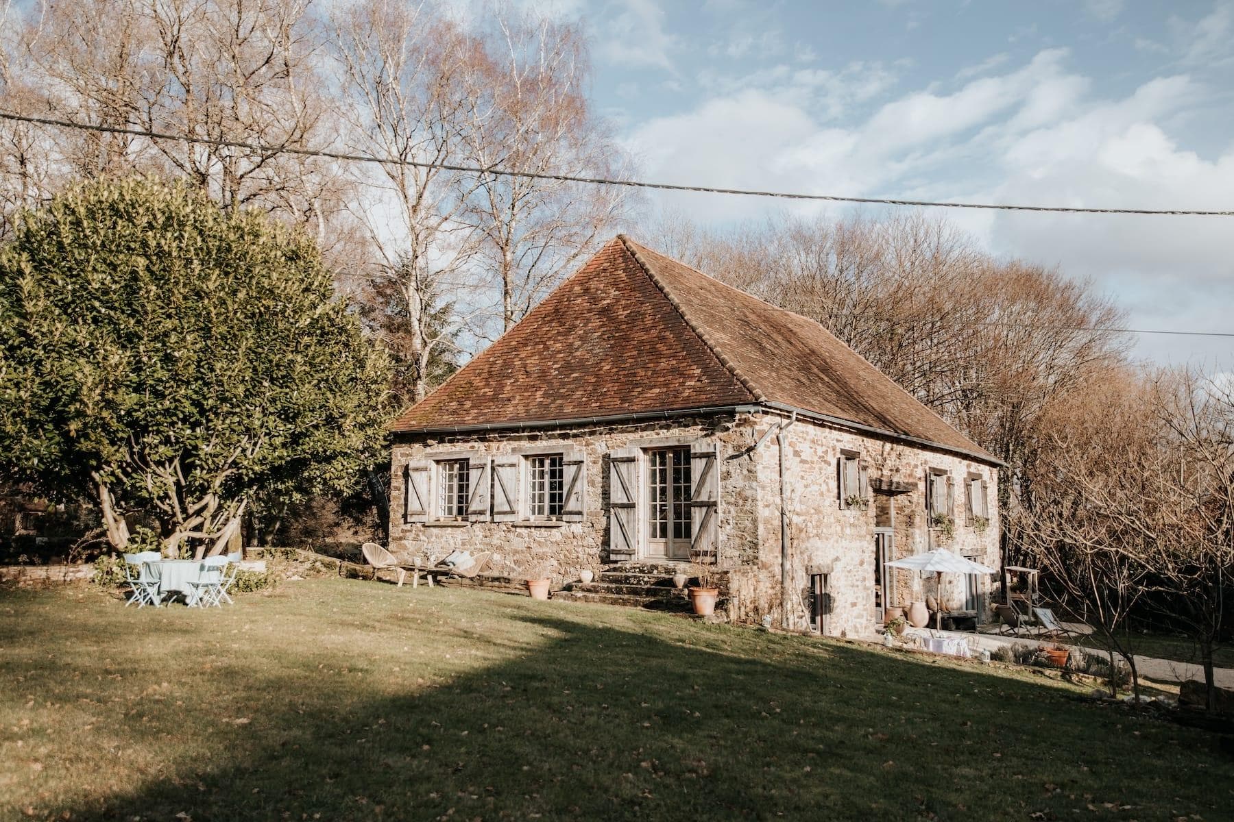 Une maison rustique en pierre avec un toit pentu en tuiles rouges est entourée d'une pelouse verte et d'arbres nus. Le soleil projette des ombres sur l'herbe. Une table de pique-nique bleue et des chaises sont installées près de la maison, et un petit auvent blanc est visible.
