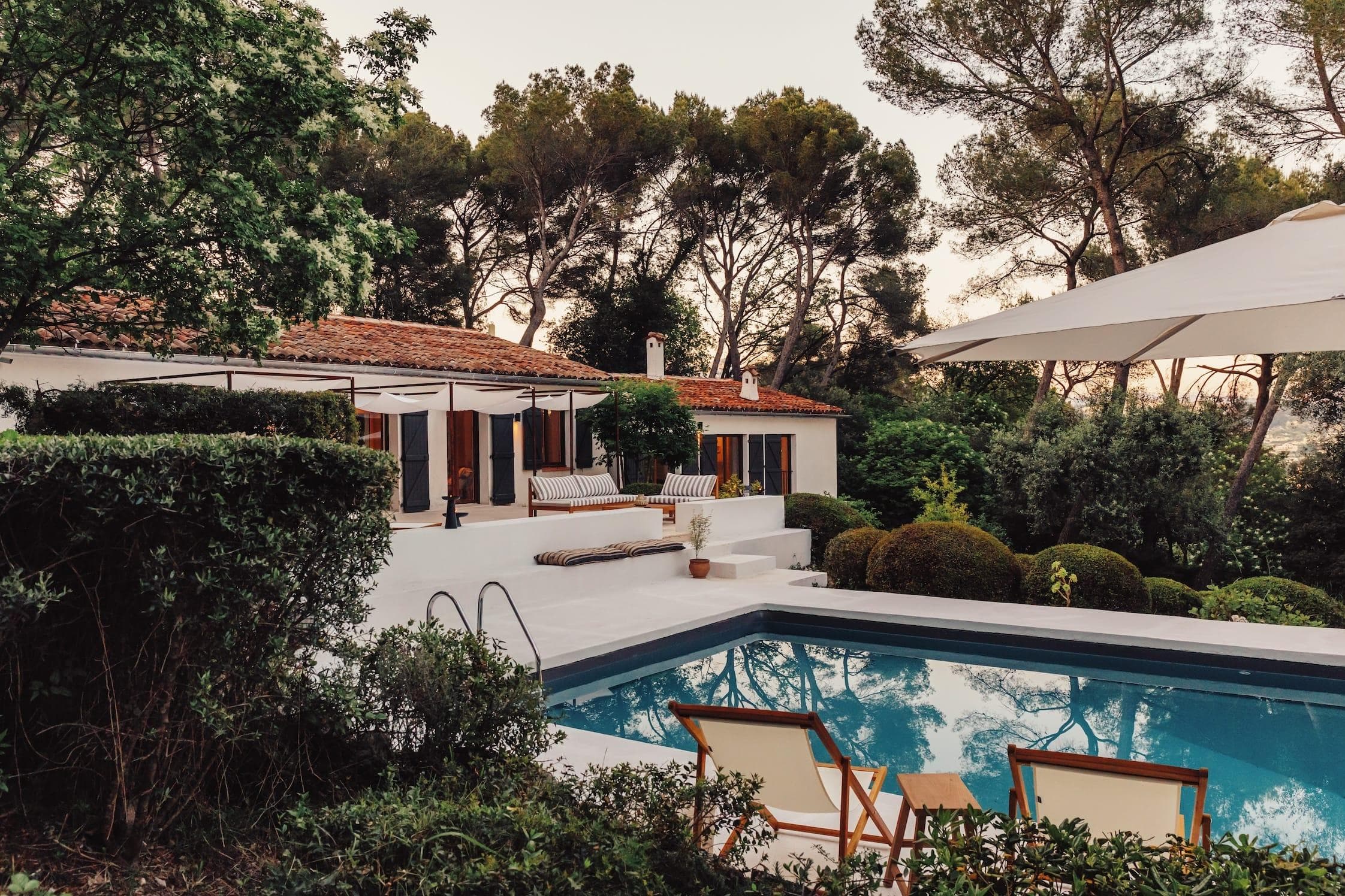 View of a single-storey house with a red tiled roof, surrounded by trees, on a sunny day. The house has white walls and large windows. In front, there is a rectangular swimming pool with a clear reflection. Garden plants can be seen all around.