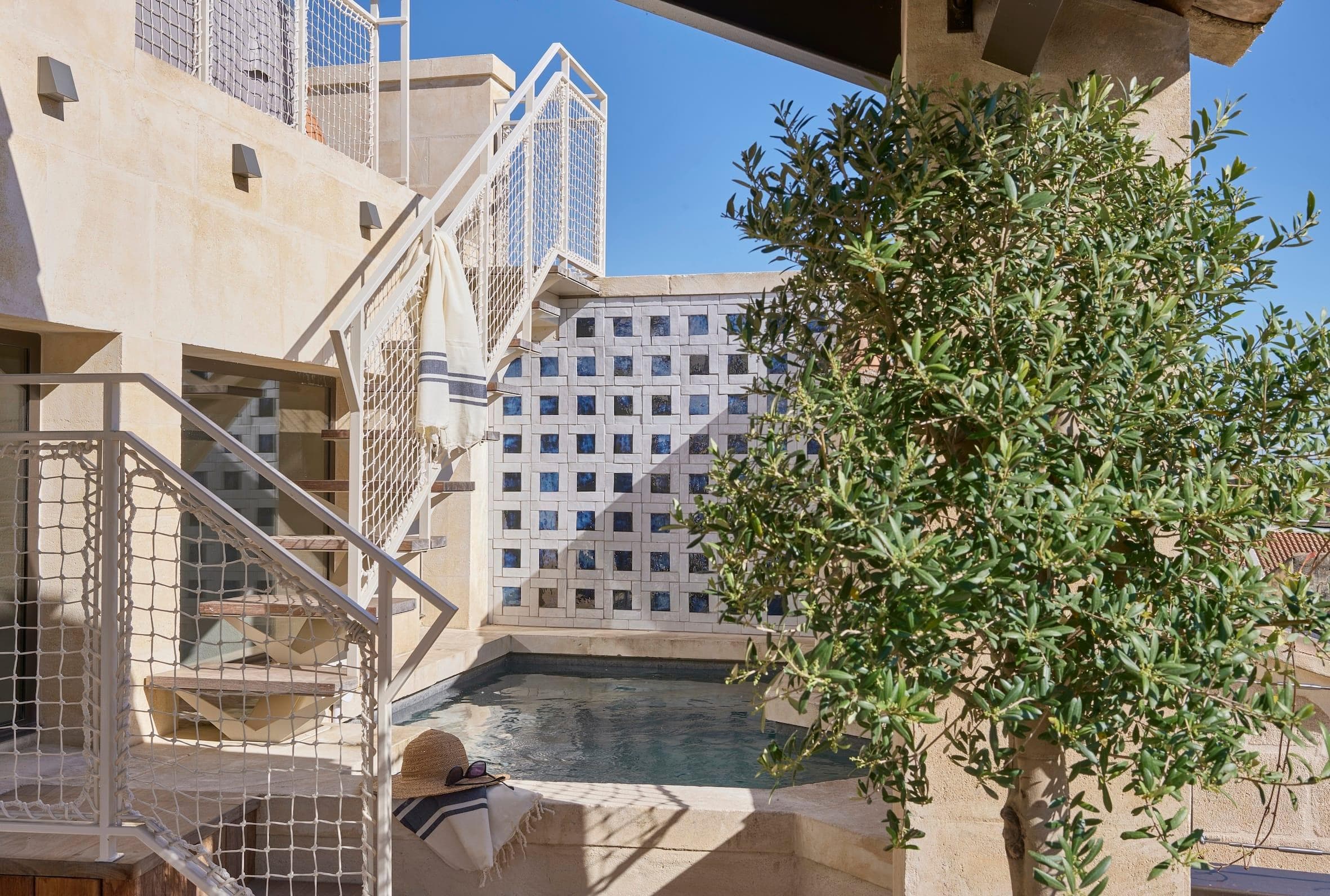 Une petite piscine extérieure est entourée de murs en pierre et de balustrades en métal blanc, avec un arbre vert feuillu au premier plan et un mur à motifs à l'arrière-plan sous un ciel bleu clair.