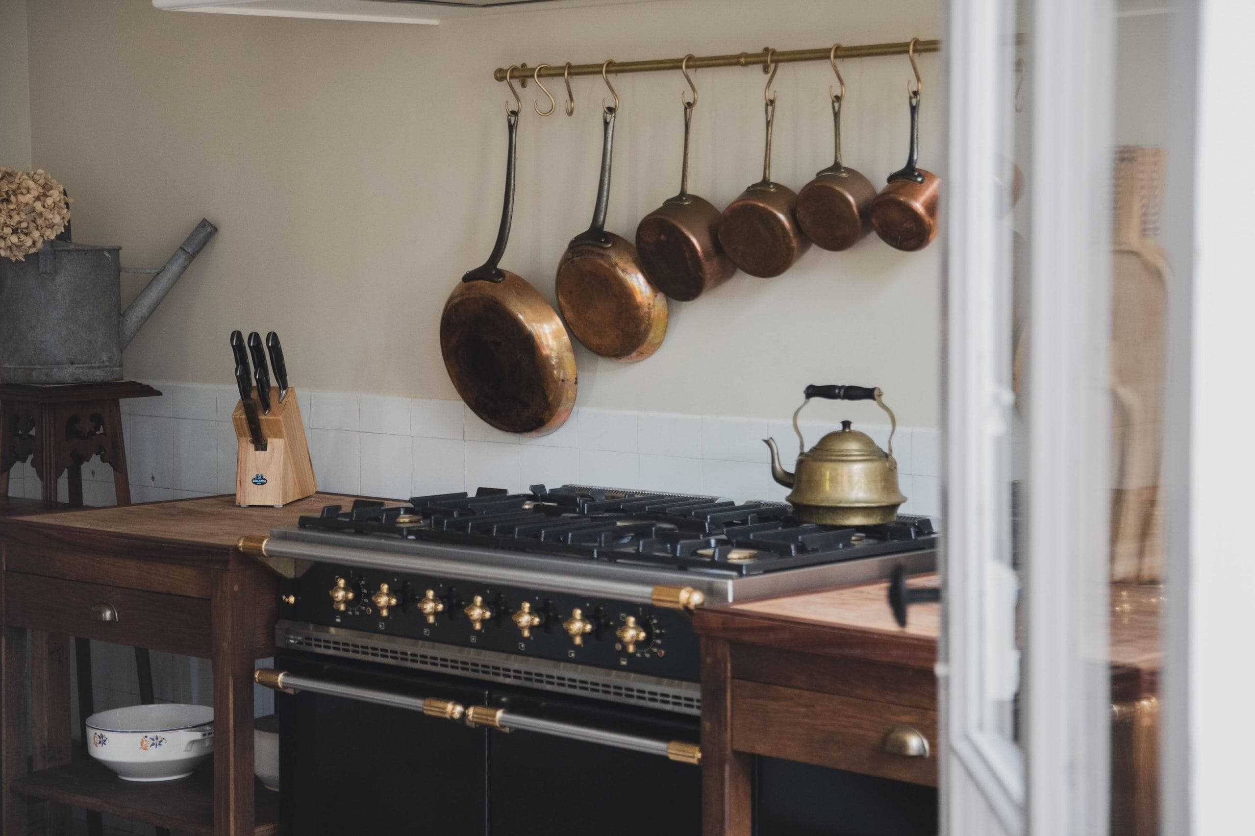 Une cuisine avec des plans de travail en bois comprend une cuisinière noire avec une bouilloire en laiton. Au-dessus de la cuisinière, plusieurs casseroles en cuivre sont suspendues à une tige. Un bloc à couteaux avec des couteaux est posé sur le comptoir, et la pièce dégage une atmosphère rustique et chaleureuse.