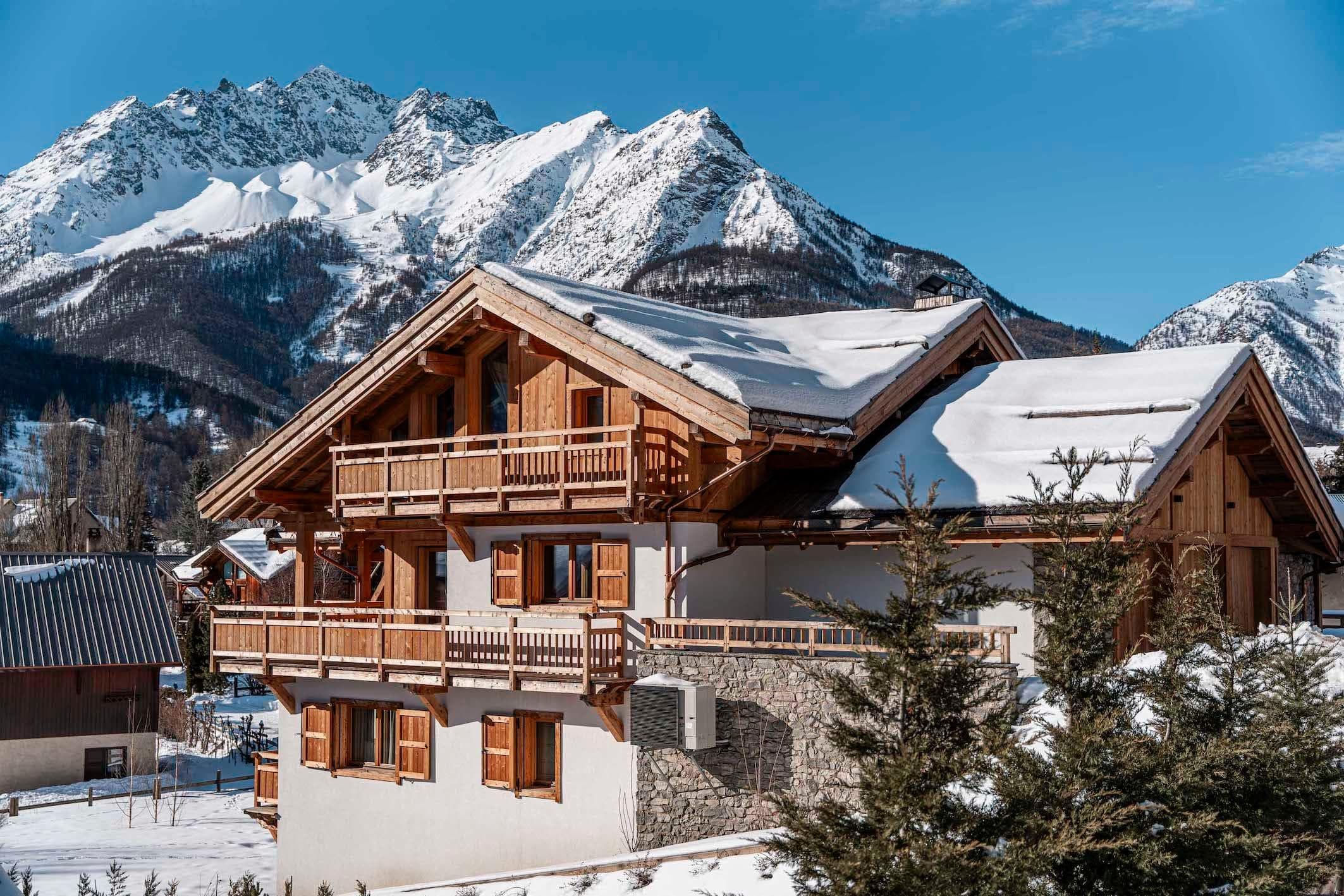 Un chalet en bois au toit enneigé se dresse sur un fond de majestueuses montagnes enneigées sous un ciel bleu limpide. Des pins sont visibles au premier plan.