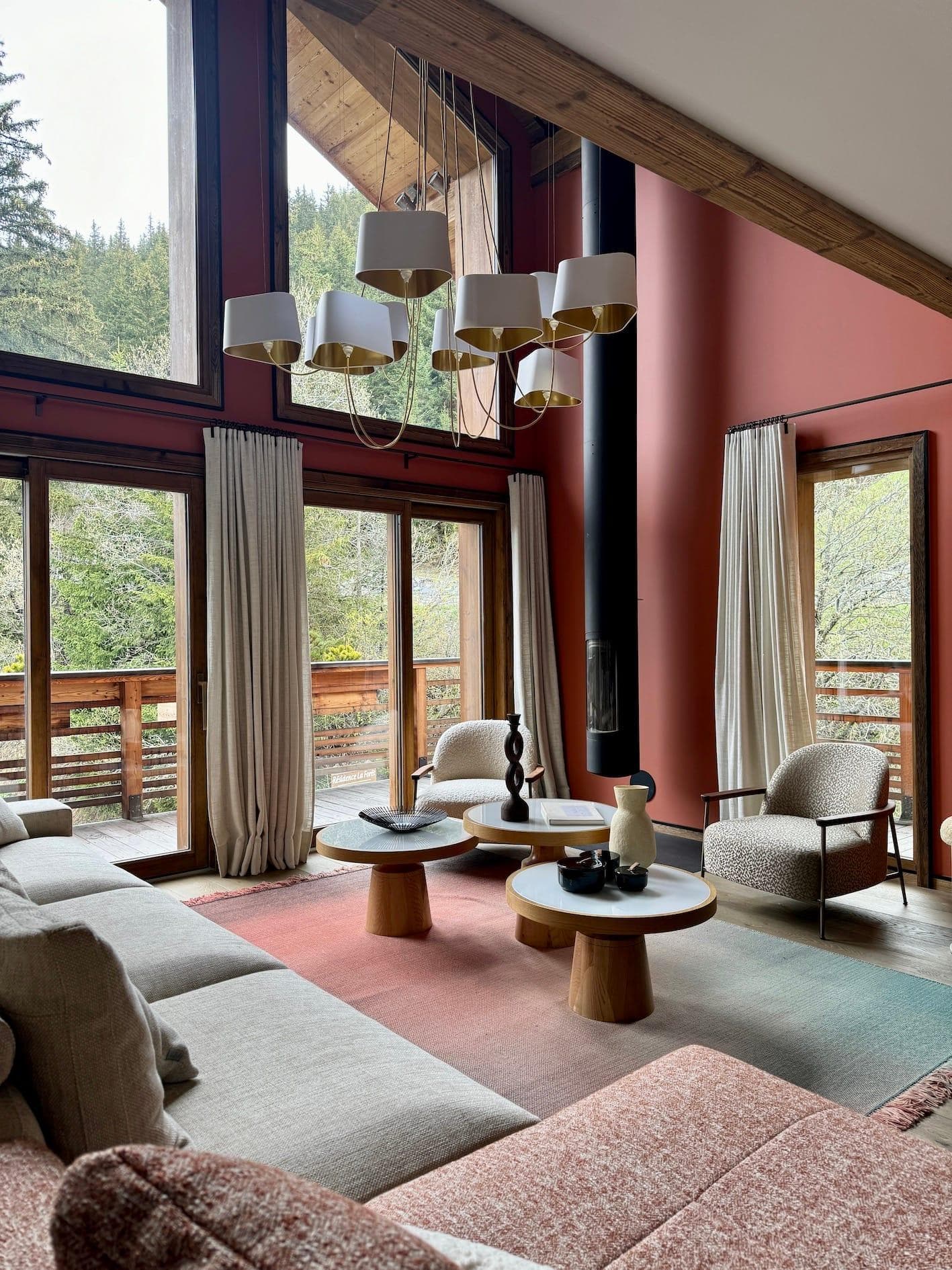 A cozy living room with large windows, high ceilings, and a view of trees. It features a sectional sofa, two round coffee tables, an armchair, and a modern chandelier. Light-colored curtains and a red accent wall add warmth to the space.