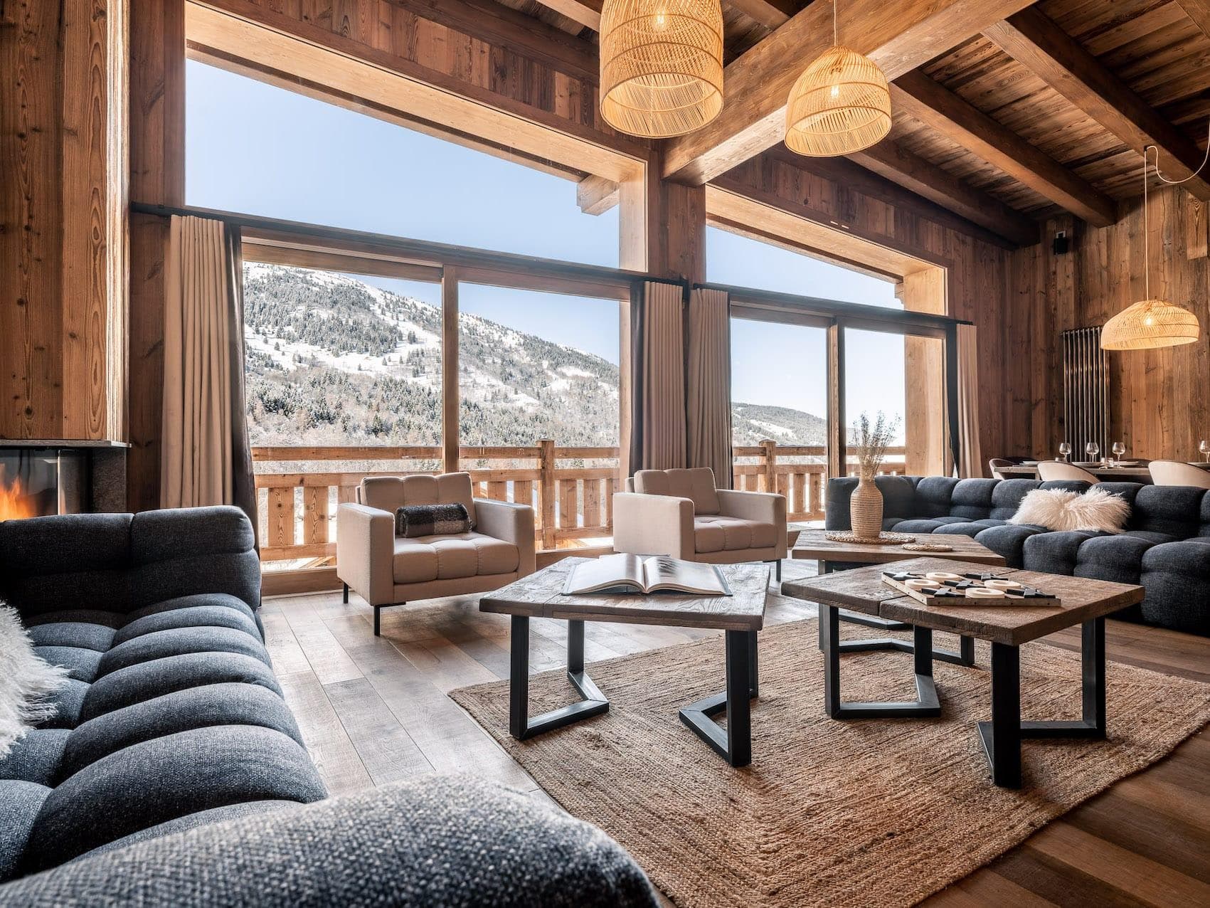 Salon spacieux avec de grandes fenêtres offrant une vue sur un paysage de montagnes enneigées. La pièce dispose de canapés sombres et clairs, d'un plafond en bois et de luminaires suspendus. Une table basse avec des livres et un tapis tissé complètent l'atmosphère chaleureuse.