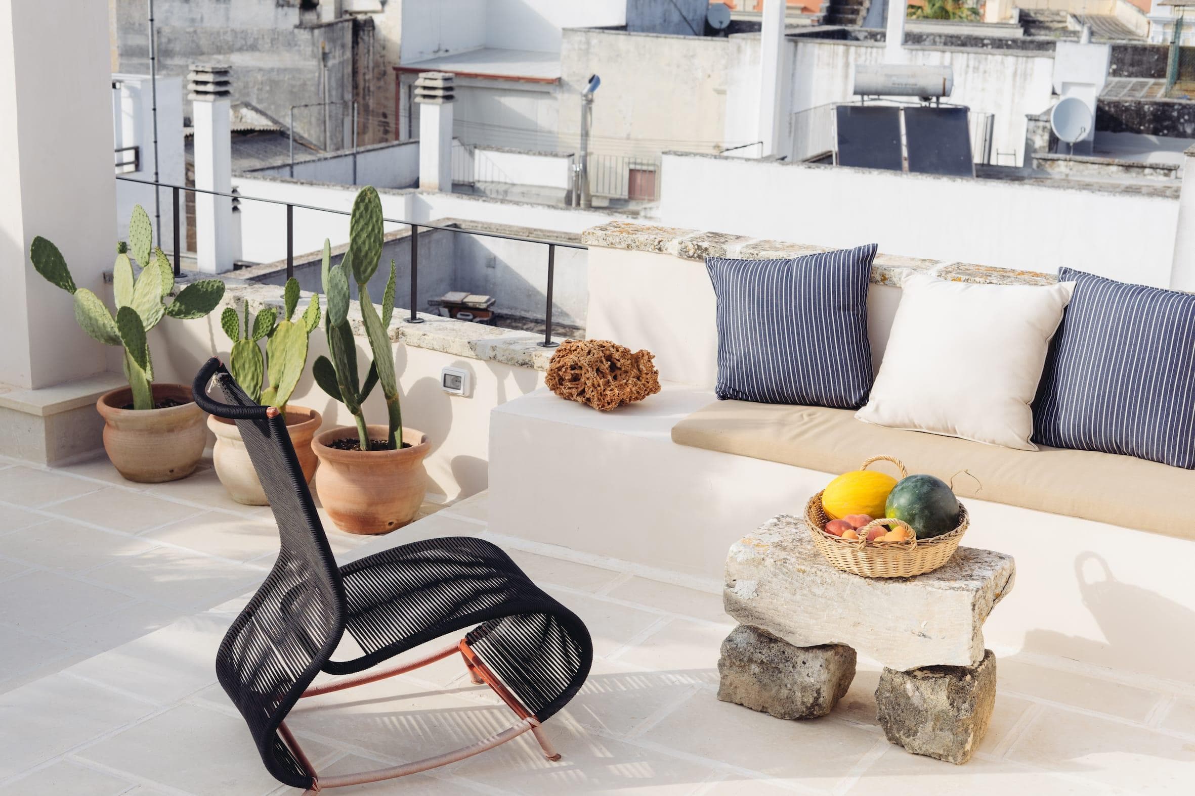 Terrasse sur le toit avec une chaise noire, une table en pierre et un banc intégré orné de coussins rayés et unis. Des cactus en pot bordent la zone et un panier de fruits est posé sur la table. Des bâtiments urbains sont visibles en arrière-plan.
