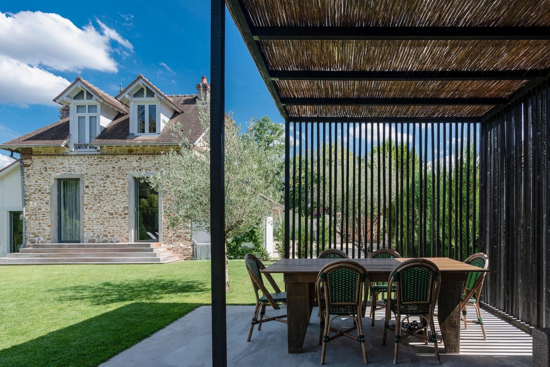 Une maison rustique avec une façade en pierre se dresse sur une pelouse bien entretenue. Au premier plan, il y a un coin repas extérieur ombragé avec une table et des chaises en bois sous une pergola en lattes sombres. Le ciel est clair avec quelques nuages duveteux.