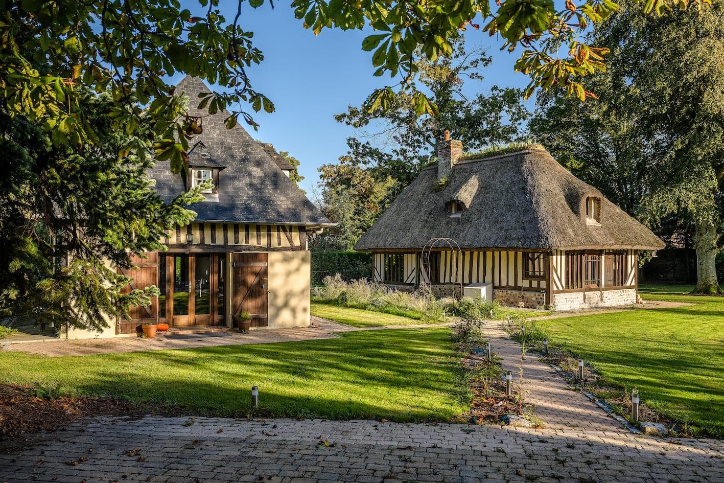 A picturesque scene with two traditional thatched-roof cottages surrounded by lush greenery. A stone pathway leads between them, and sunlight filters through the trees, casting gentle shadows on the grass.