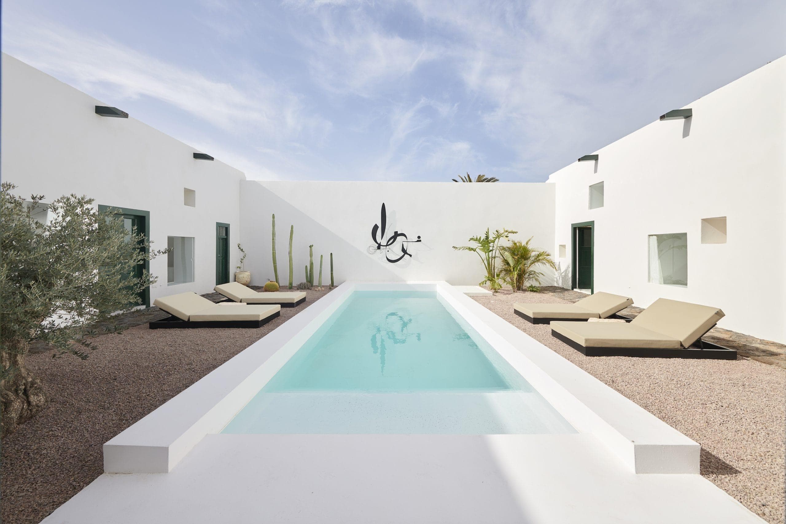 Modern minimalist courtyard with a central rectangular swimming pool, surrounded by beige lounge chairs, potted cacti, and a few small trees. The white walls feature a black abstract sculpture. Blue sky with light clouds above.