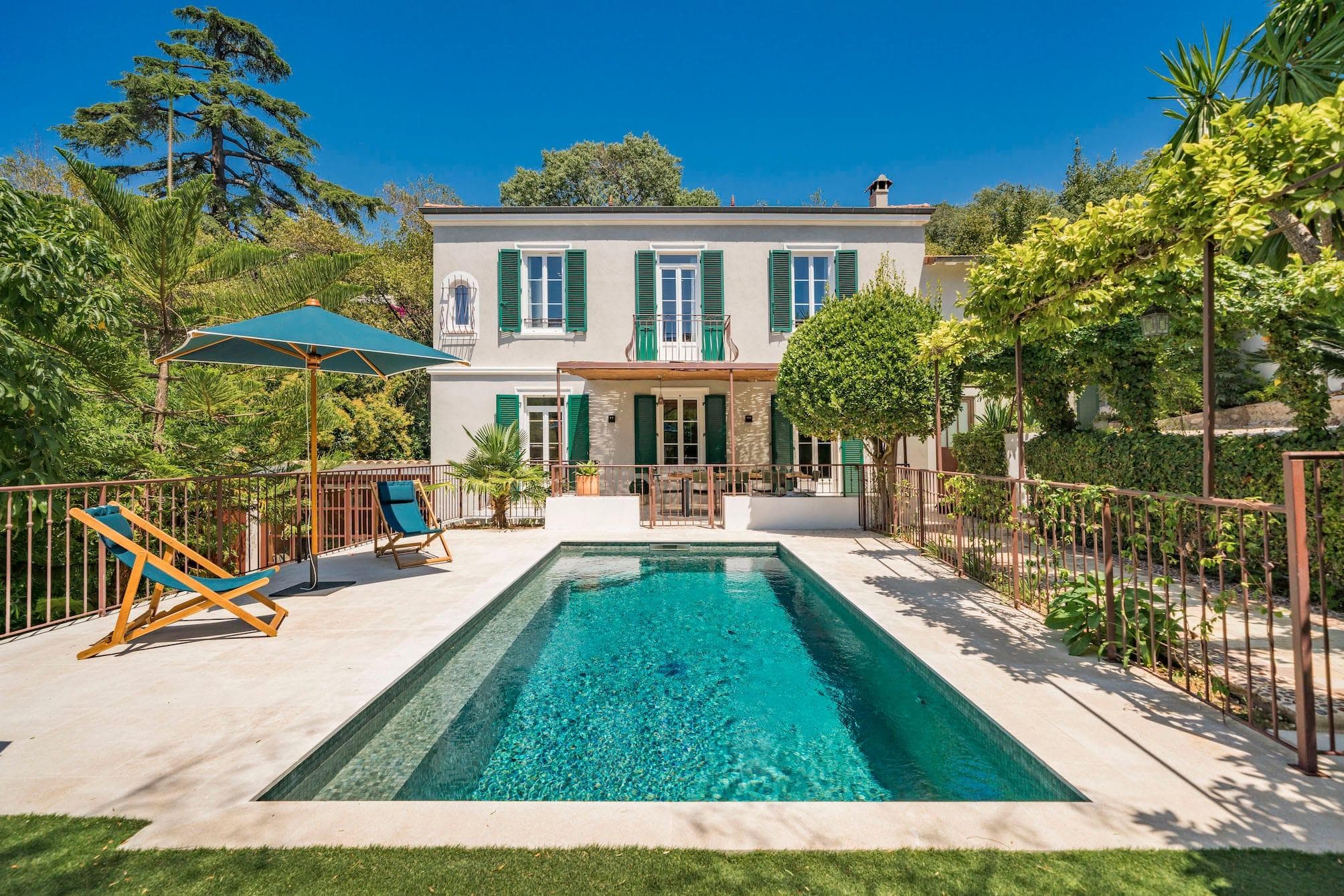 A charming two-story house with green shutters overlooks a rectangular pool. Surrounded by greenery, it features a patio with seating and an umbrella. A deck chair is beside the pool under a clear blue sky.