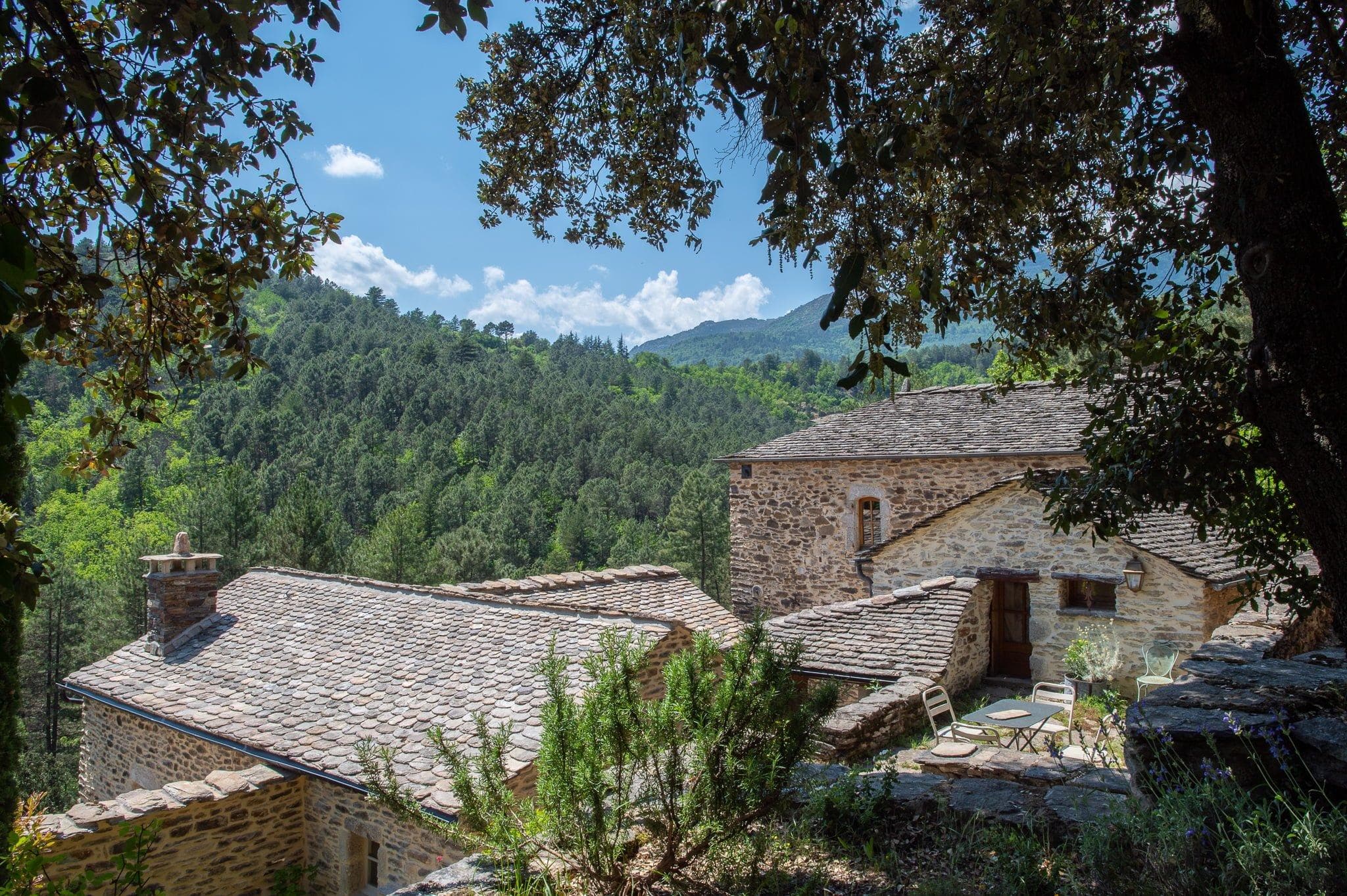 Une maison rustique en pierre avec un toit en bardeaux est nichée au milieu d'arbres verts luxuriants à flanc de montagne. La maison dispose d'espaces extérieurs pour s'asseoir et est entourée d'une forêt dense et de collines vallonnées au loin sous un ciel bleu parsemé de nuages.