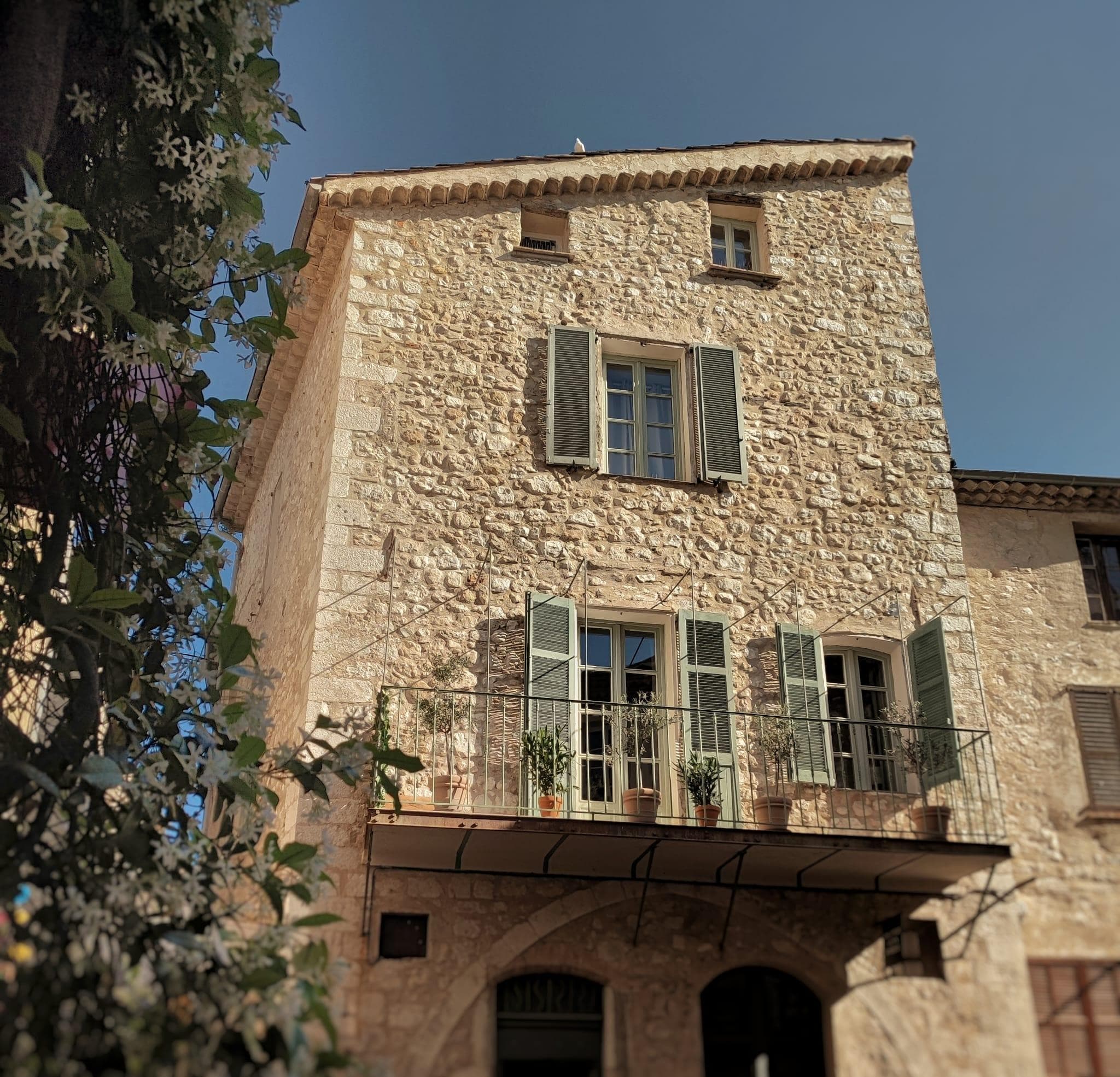 Un bâtiment rustique en pierre avec des volets verts et un petit balcon orné de plantes en pot. La façade est ensoleillée et le ciel est dégagé. Au premier plan, un arbre aux fleurs blanches apporte une touche de nature.