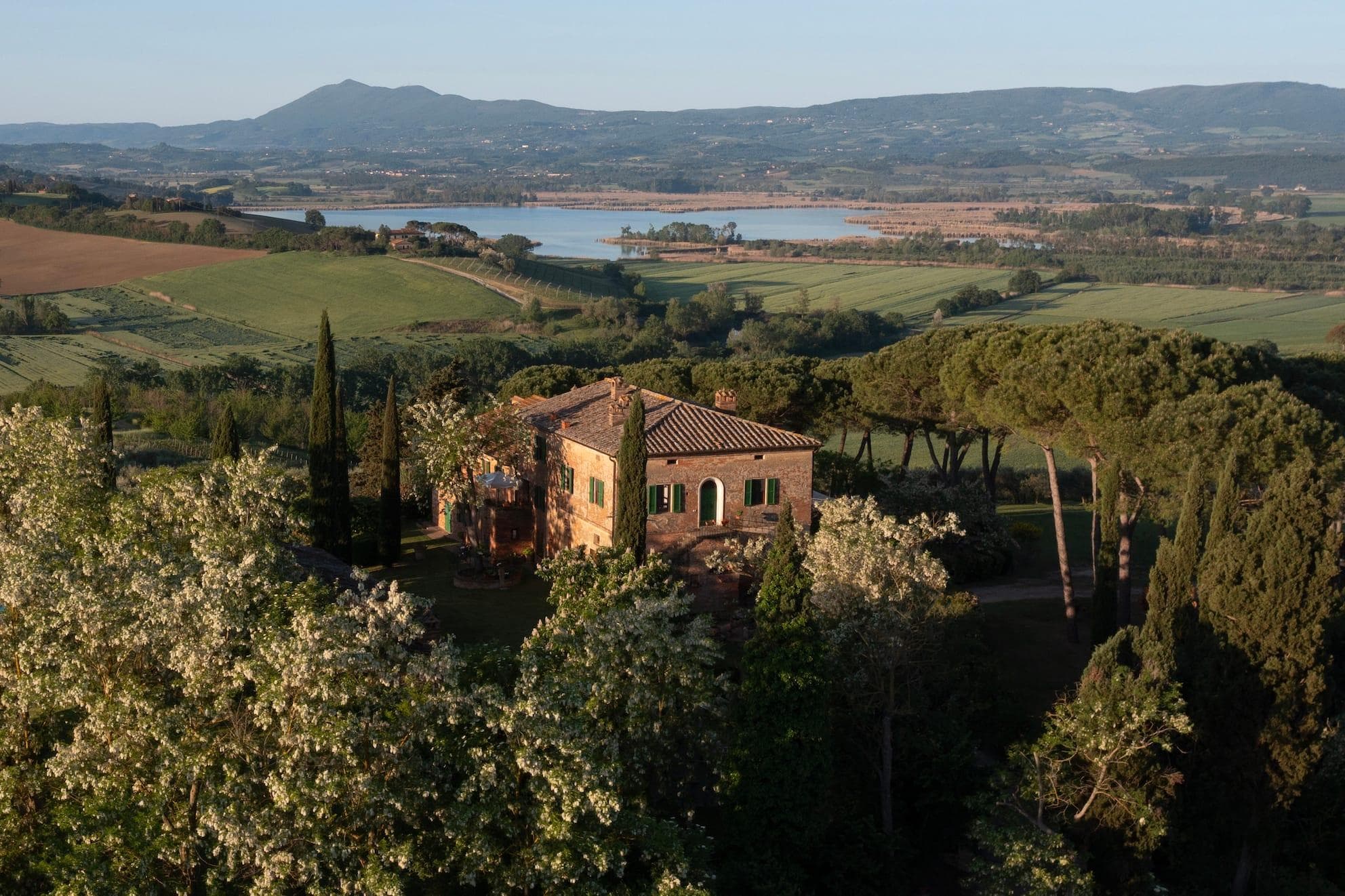 Une villa rustique entourée d'arbres luxuriants et de verdure, située dans un paysage pittoresque avec des collines ondulantes. Un lac est visible au loin, sous un ciel bleu clair avec des montagnes au loin.