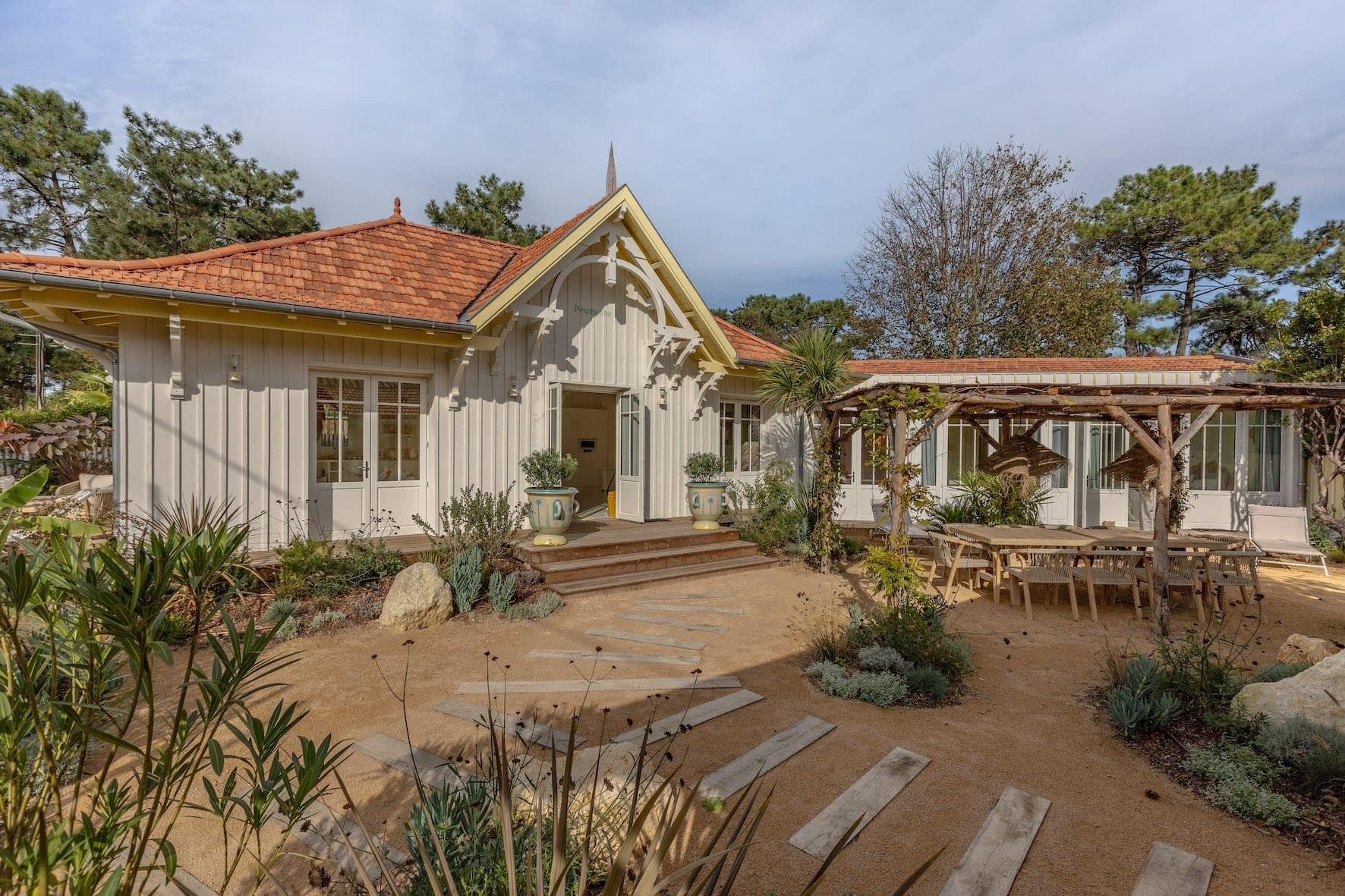 Charming cottage with beige wood cladding and red tile roof, surrounded by lush vegetation and a sandy garden. A path leads to the entrance, and a pergola-covered patio with outdoor seating is visible on the right.
