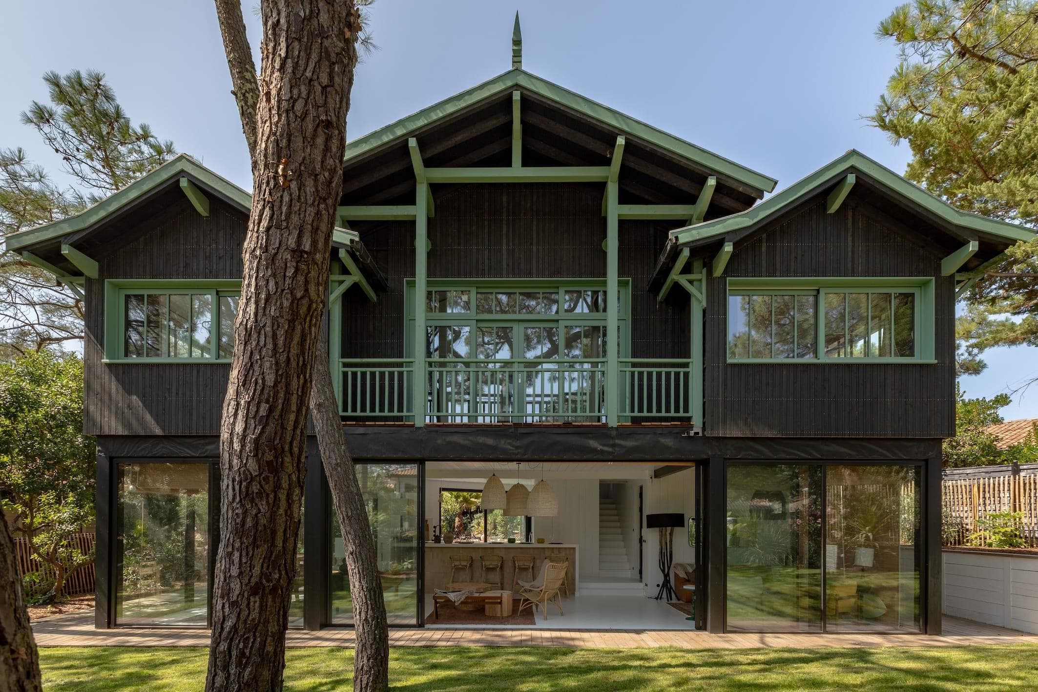 A two-story house with dark wood and green trim, featuring large windows and a balcony. It has a modern design with a spacious, open interior visible through the glass. Trees surround the house, and a well-kept lawn is in the foreground.