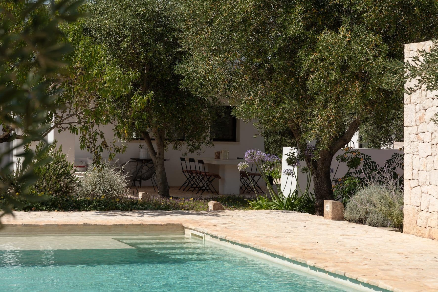 A serene outdoor scene with a swimming pool in the foreground and a rustic stone patio surrounded by trees. Several chairs are placed around a table on the patio, creating an inviting and peaceful atmosphere.