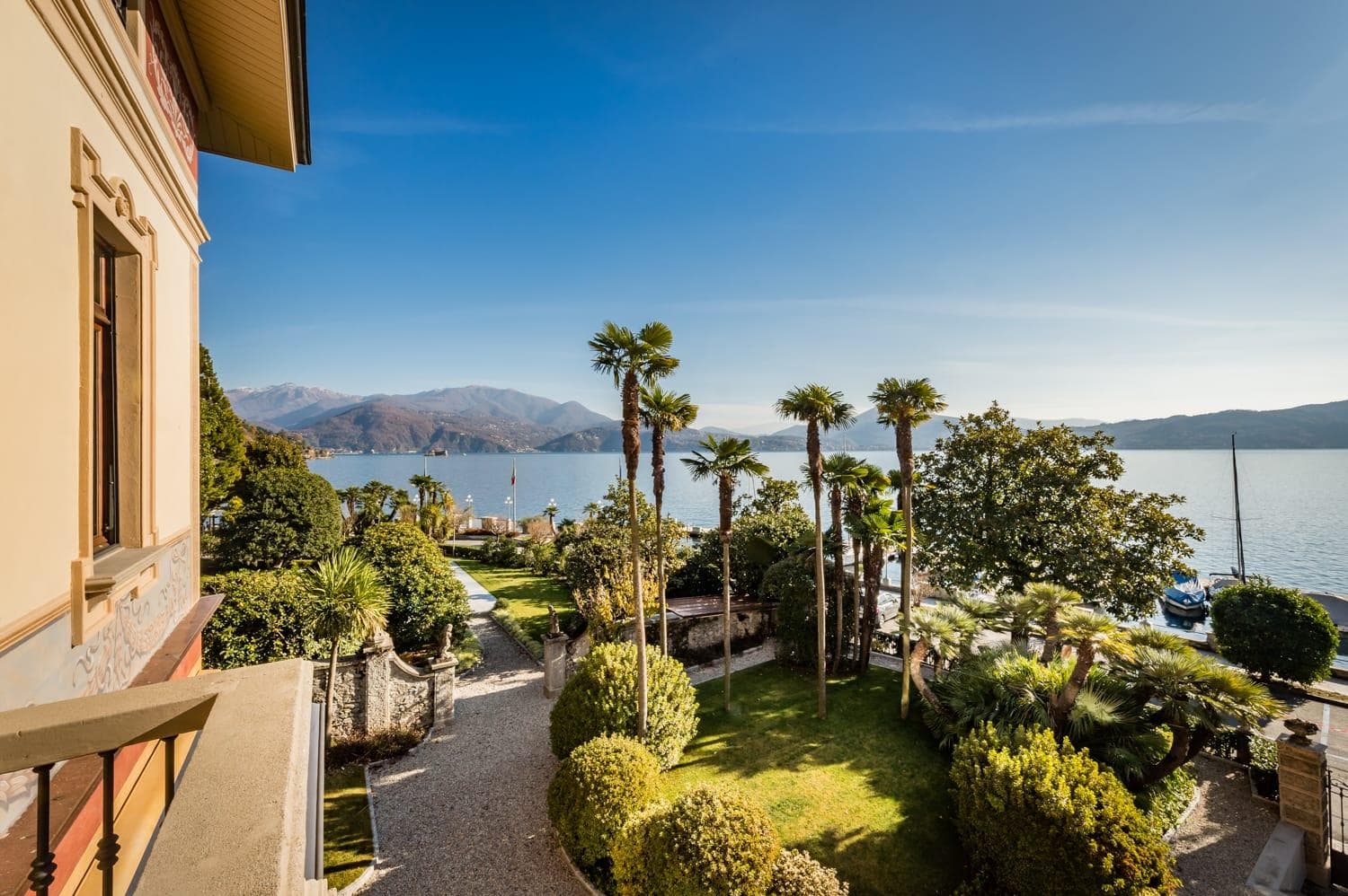 Vue depuis un balcon surplombant un lac paisible avec des montagnes au loin. Au premier plan, un jardin planté de palmiers et d'une végétation luxuriante. Un sentier mène au lac, où des bateaux sont amarrés près de la rive. Le ciel est clair et bleu.