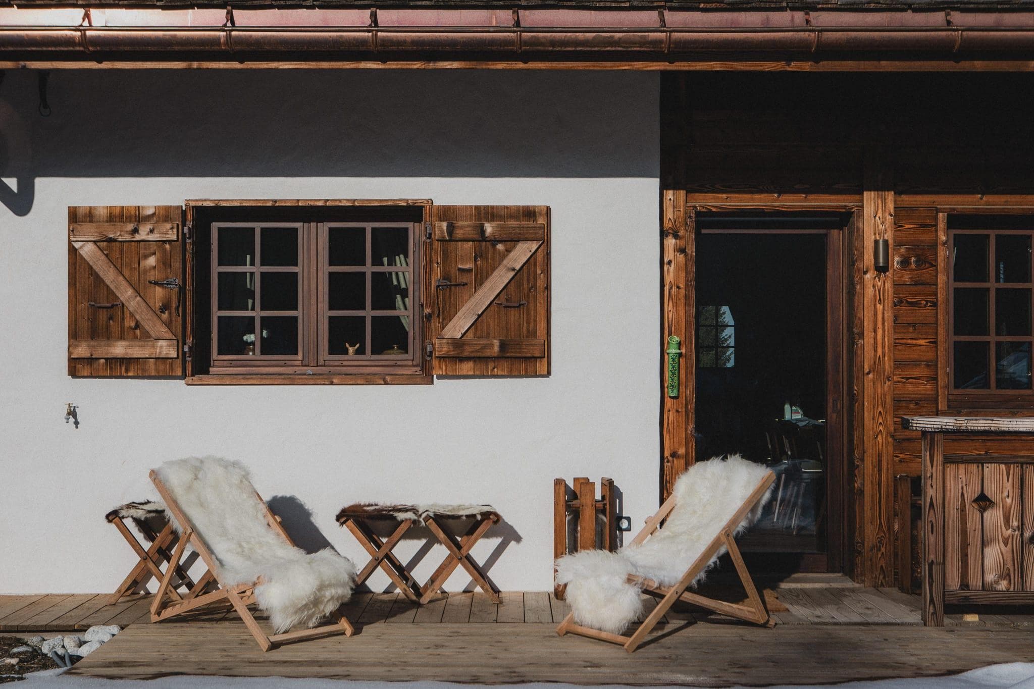 Deux chaises longues en bois recouvertes de fausse fourrure blanche sont installées sur une terrasse en bois à l'extérieur d'une maison rustique. Une petite table en bois les sépare. La maison est dotée de volets en bois, d'une porte ouverte et la lumière du soleil projette des ombres sur la terrasse.