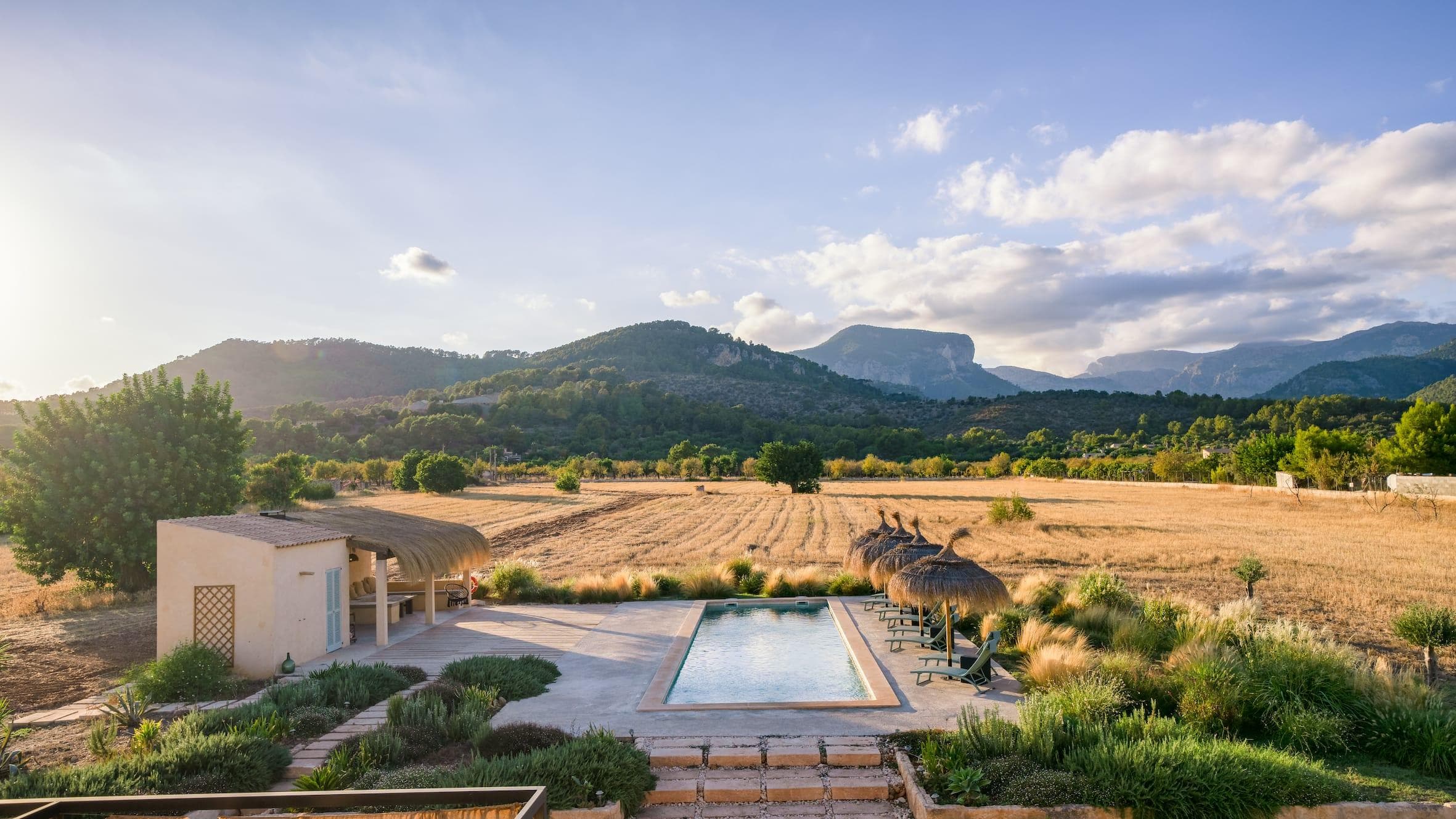 A picturesque rural landscape with a small rectangular pool surrounded by grassy fields and lush greenery. Straw umbrellas and lounge chairs line one side of the pool. Mountains and a partly cloudy sky are visible in the background.