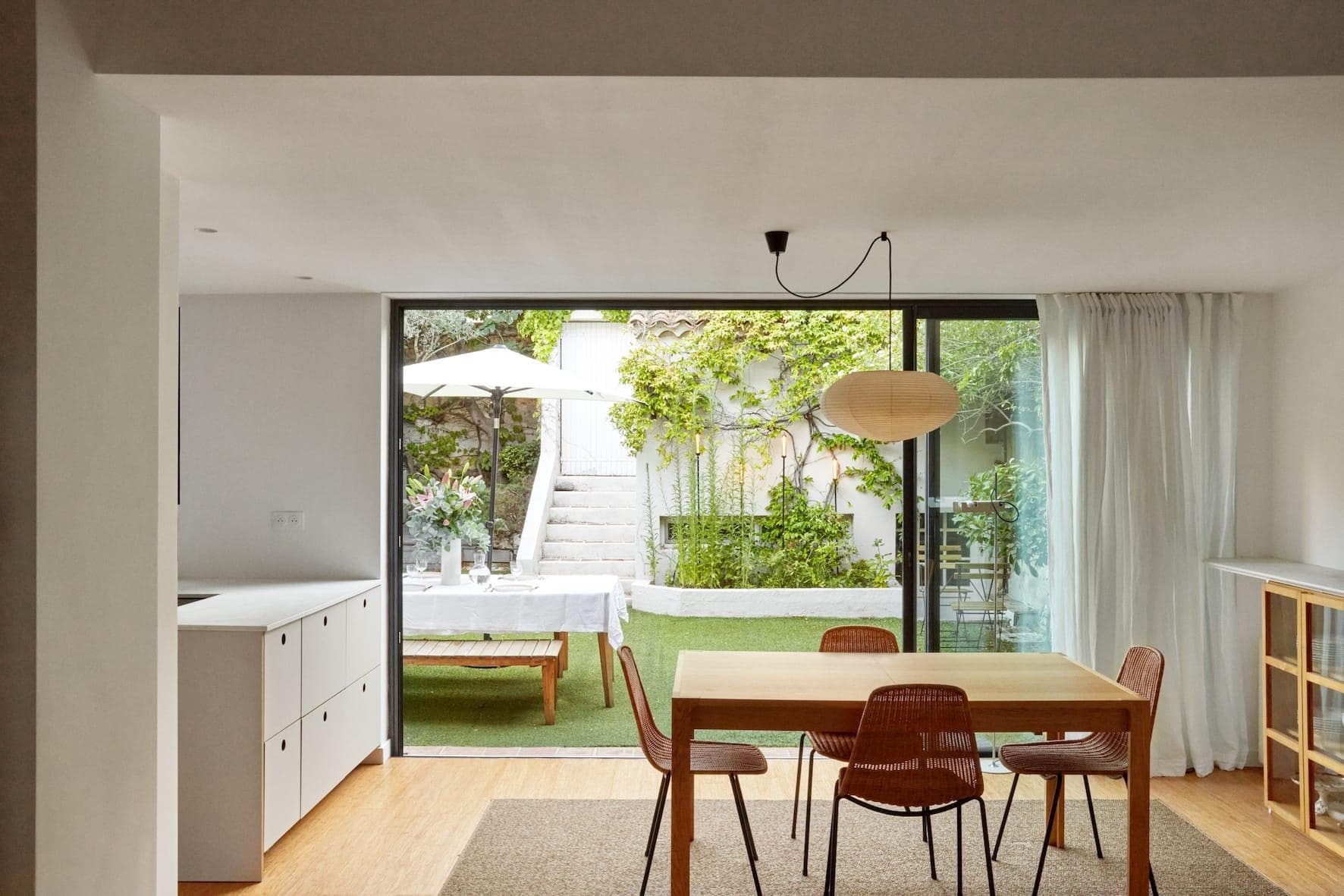 Salle à manger minimaliste avec une table en bois et quatre chaises posées sur un tapis. De grandes portes vitrées coulissantes s'ouvrent sur un jardin luxuriant agrémenté de plantes, d'une table blanche, de chaises et d'un parasol. Un éclairage tamisé et des tons naturels créent une atmosphère sereine.