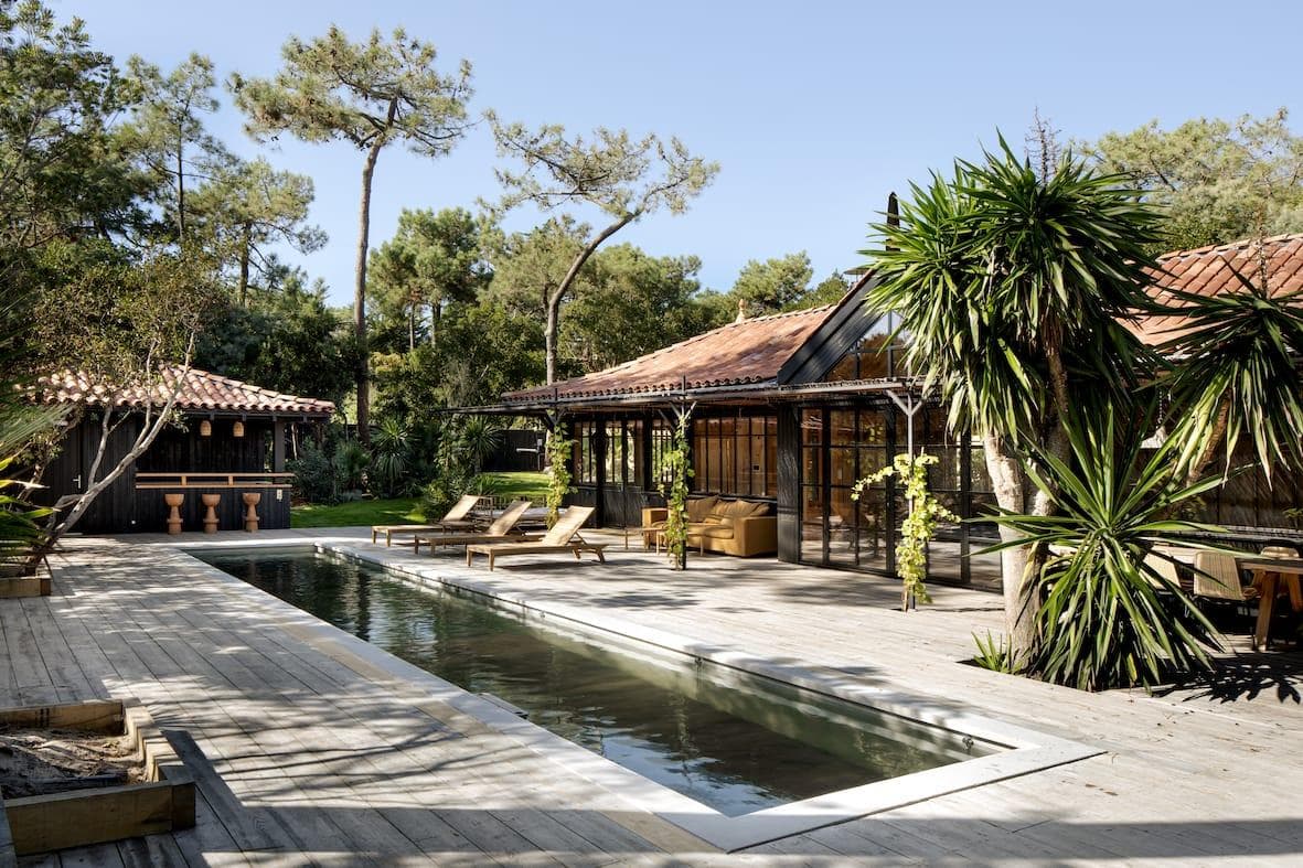Luxurious outdoor pool area with wooden decking, surrounded by lush greenery. The setting includes lounge chairs, a pergola, and a building with a tiled roof. Tall trees are visible in the background under a clear sky.