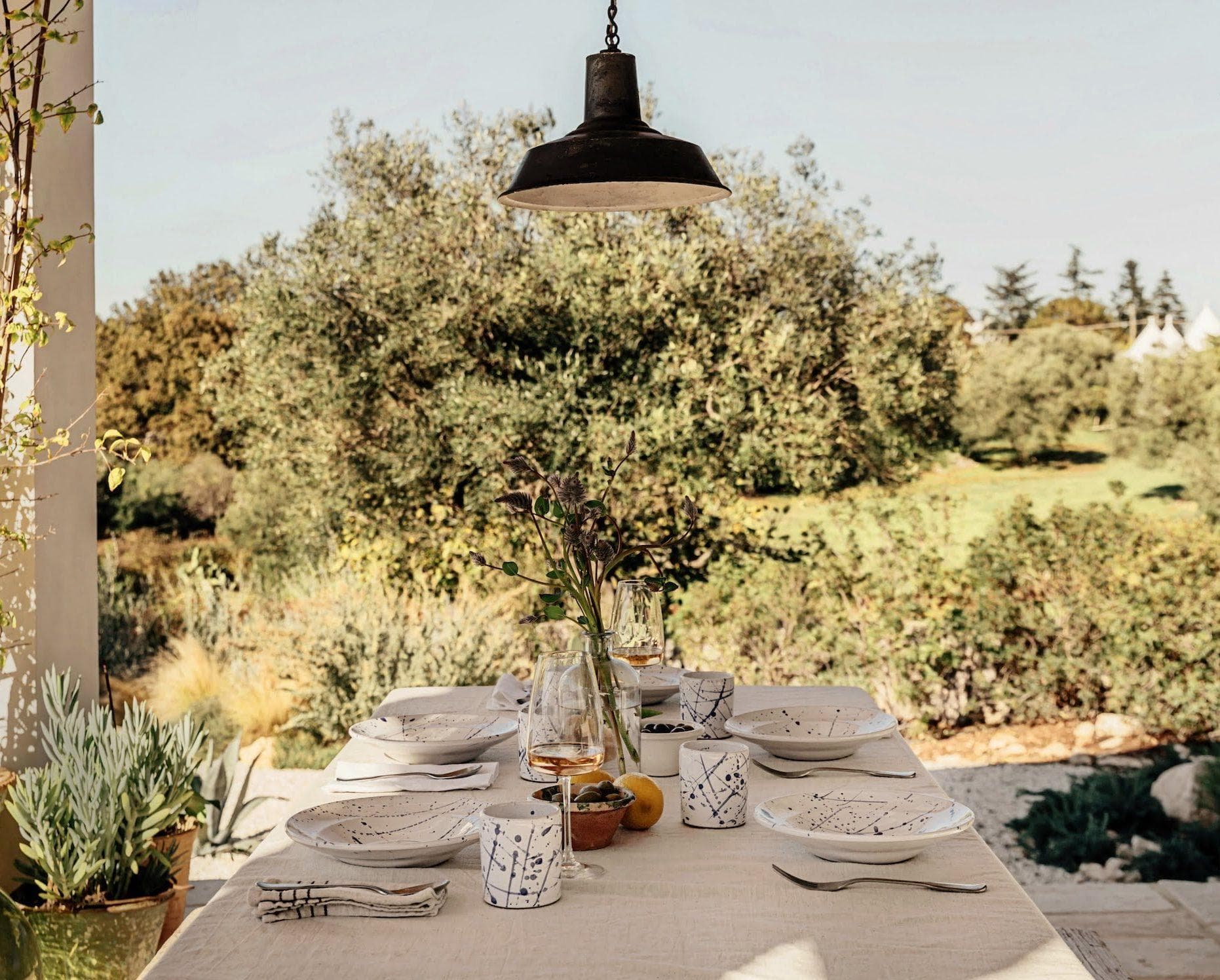 Une table à manger d'extérieur joliment dressée avec du linge blanc, de la vaisselle et un centre de table composé de fleurs, placée sous une lampe suspendue. L'arrière-plan présente des arbres et des plantes vertes luxuriantes, créant une atmosphère sereine et accueillante pour un repas.