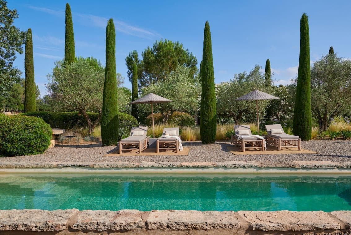 Three lounge chairs with umbrellas are set on a gravel patio beside a turquoise pool, surrounded by tall cypress trees and lush greenery under a clear blue sky.