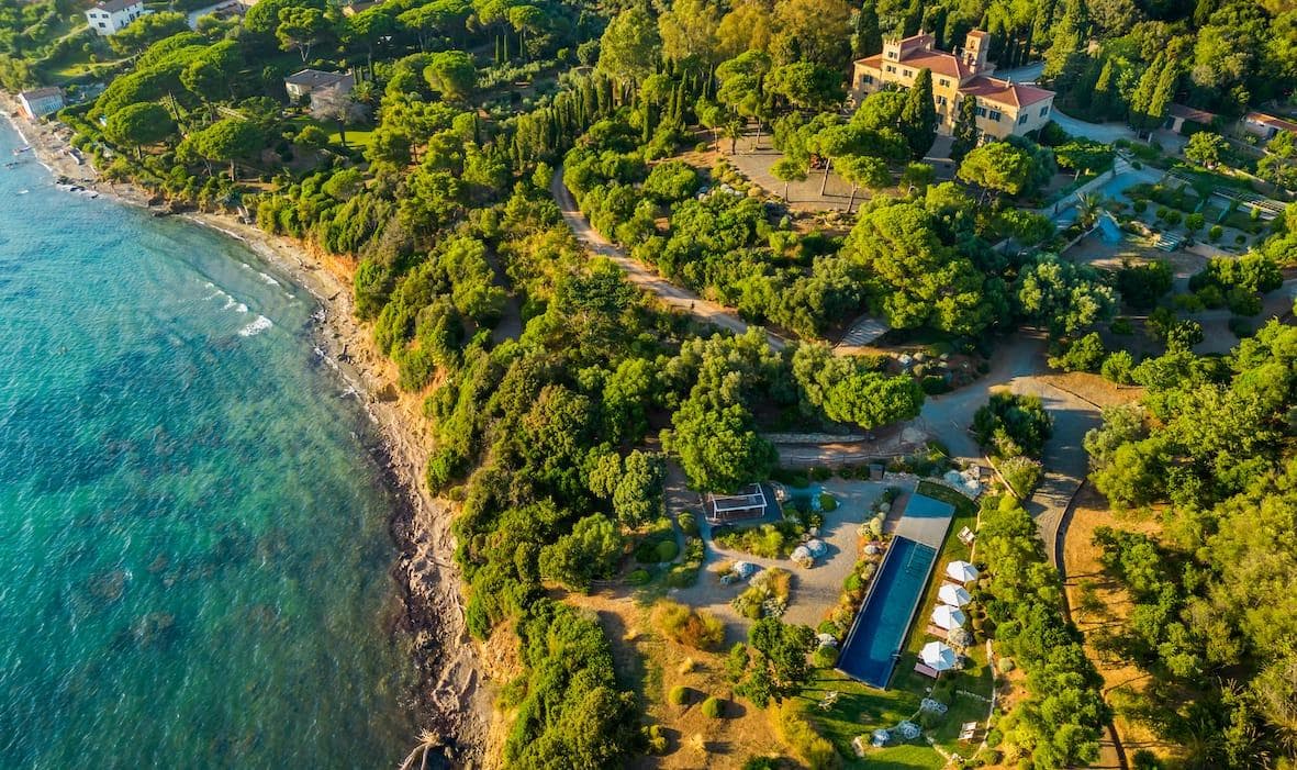 Vue aérienne d'un paysage côtier verdoyant et luxuriant, avec une maison entourée d'arbres denses. Une plage étroite aux douces vagues borde la rive gauche, tandis qu'une piscine rectangulaire avec chaises longues est visible au milieu de la verdure à droite.